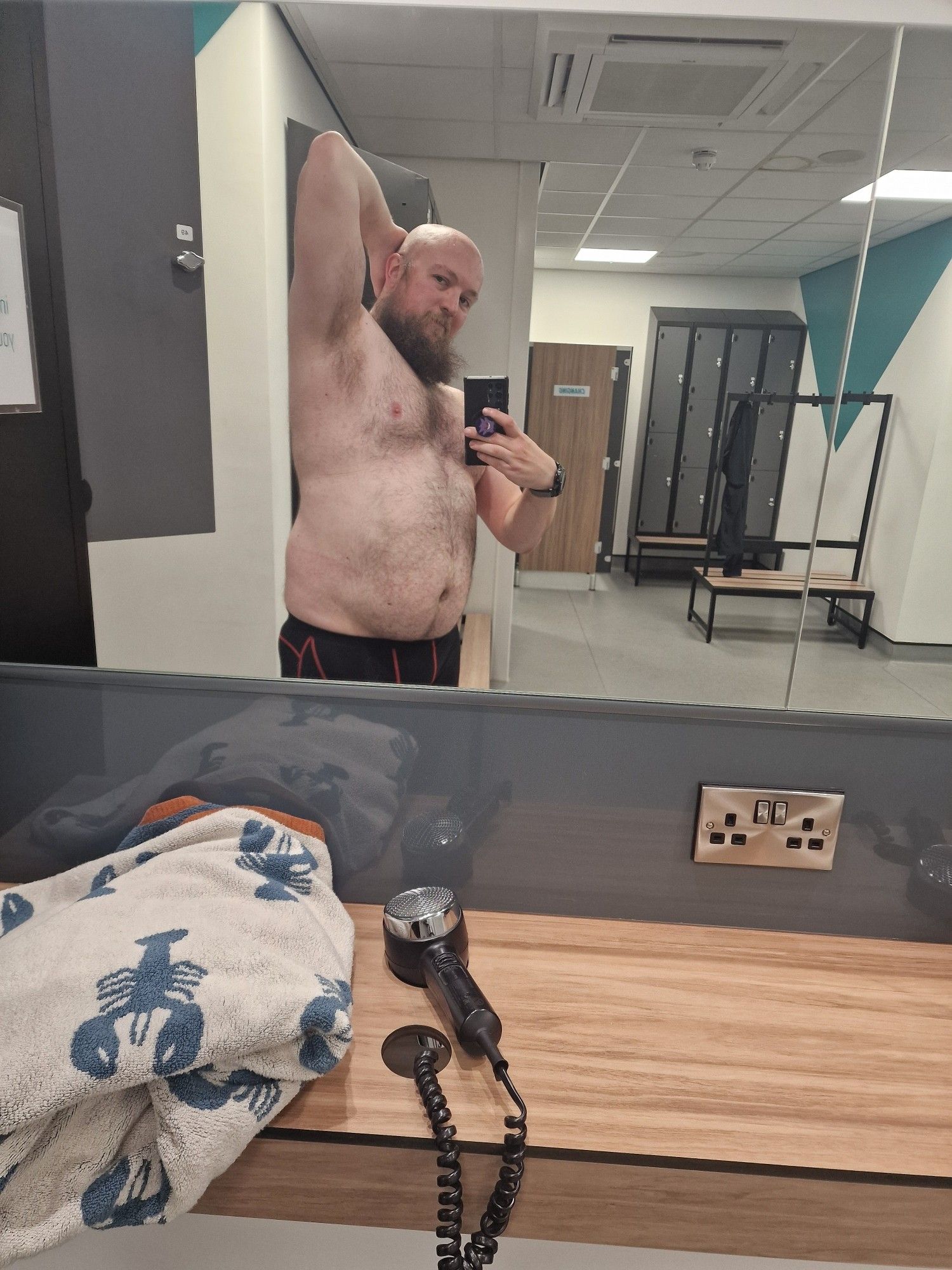 A man stands taking a photo on his phone in the reflection of a mirror in a gym changing room. The man is the main focus of the image. He is wearing only underwear and is visible in the reflection of the mirror from the waist up. He would be described as a "bear" in the gay community, is bald and has a beard that extends to his collarbone. He is has his arm extended upward with his hand behind his head. He has reasonable musculature and is overweight with a round belly.