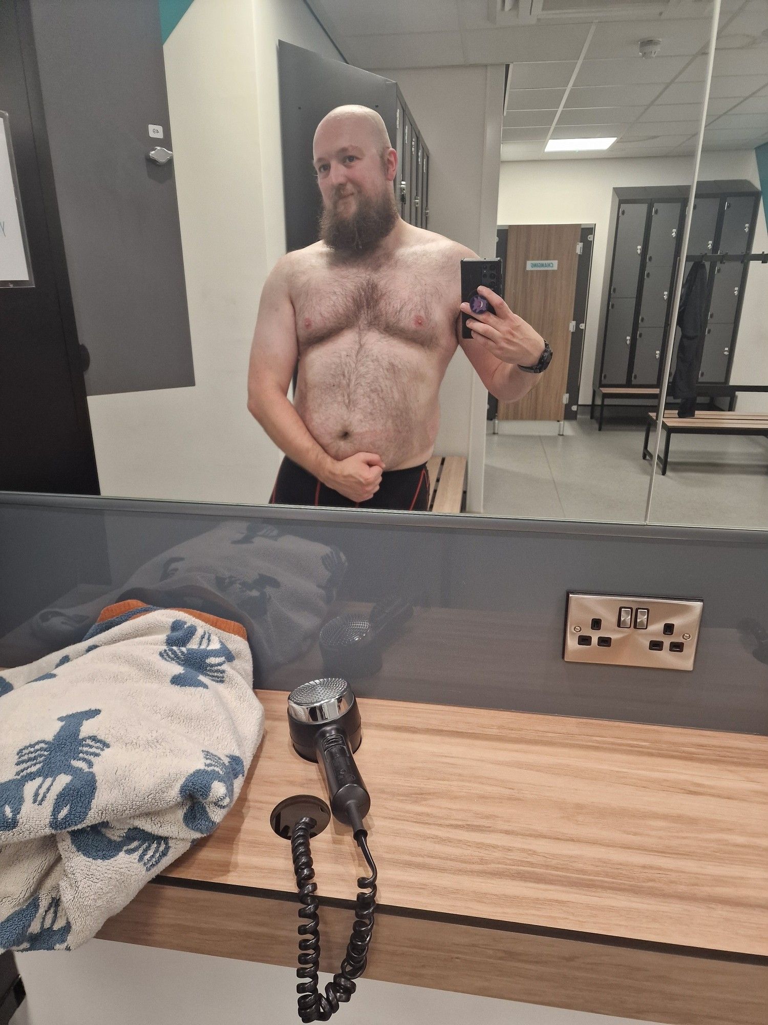 A man stands taking a photo on his phone in the reflection of a mirror in a gym changing room. The man is the main focus of the image. He is wearing only underwear and is visible in the reflection of the mirror from the waist up. He is flexing his arm in a traditional pose to accent his bicep. He has reasonable musculature and is overweight with a round belly.