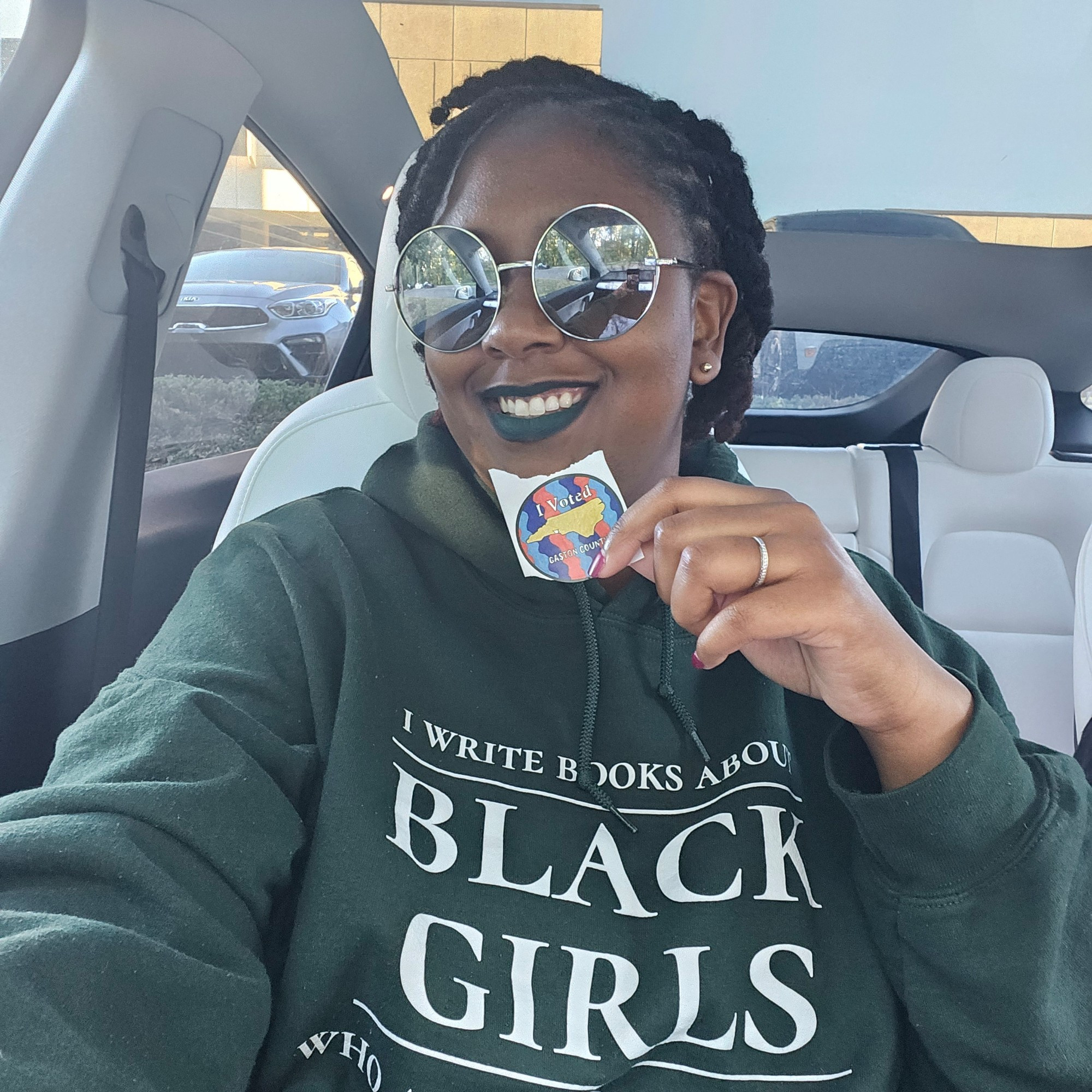 A Black woman with green lipstick, sunglasses, locs, with a green hoodie that reads: I wrote books about Black girls who aren't all that nice holds up a sticker that reads I Voted