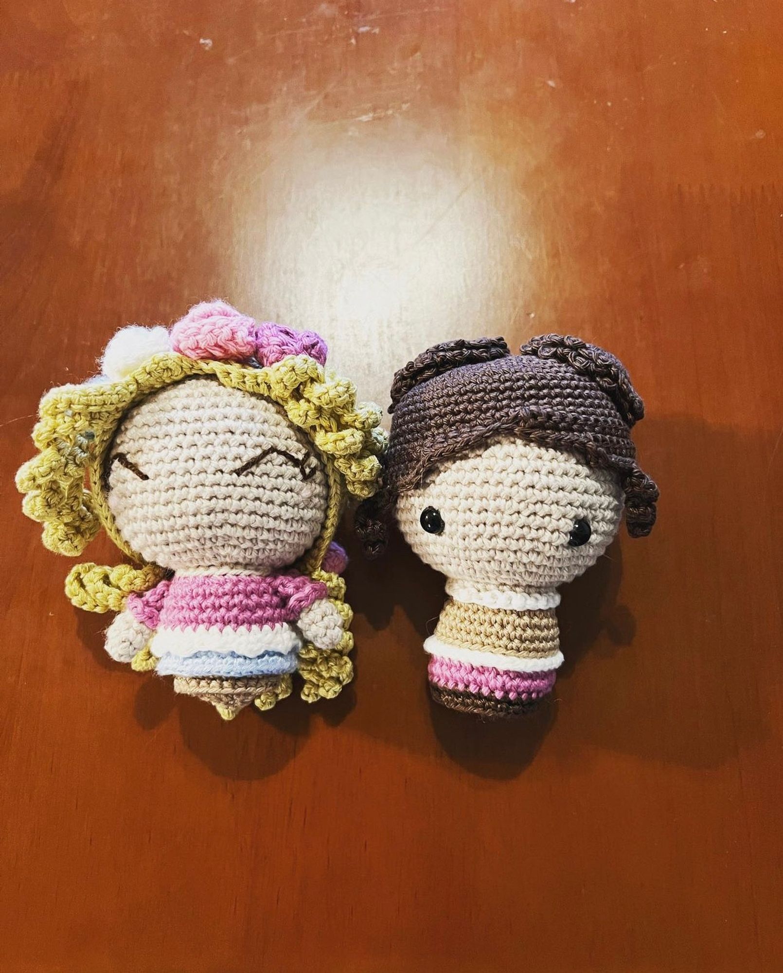 Two small crochet dolls sit on a wooden table. One has curly blonde hair, a pink shirt, and a blue skirt. The other has brown hair pulled up into 2 buns, a tan shirt, and pink skirt.