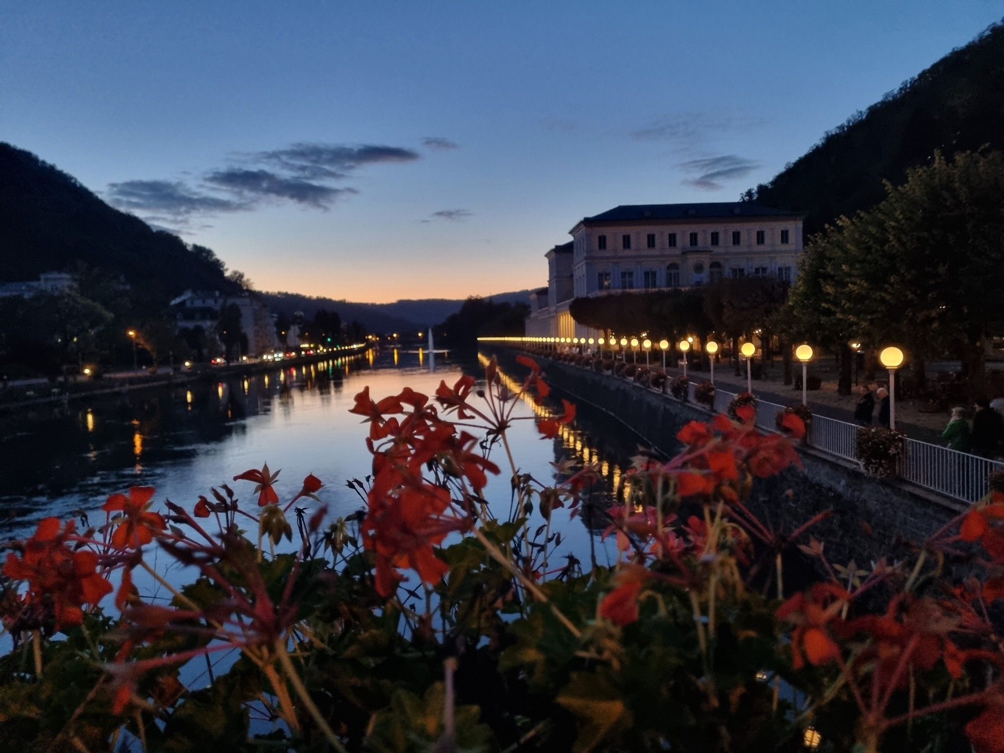 Im Vordergrund sieht man rote Geranien, dahinter zum.oberen Bildrand hin einen Fluss. Rechts eine Reihe kugelförmiger Leuchten an  einem Geländer, das sich an der Lahn entlangzieht. Zwei größere Gebäude stehen dort weiter hinten. Links sind ebenfalls Lichter zu sehen und Gebäude, allerdings liegen sie schon im Dunklen.
Der Himmel ist hellblau, mit kleinen grauen Wolken und einem rosafarbenen Streifen am Horizont. An den Seiten sieht man Hügel.