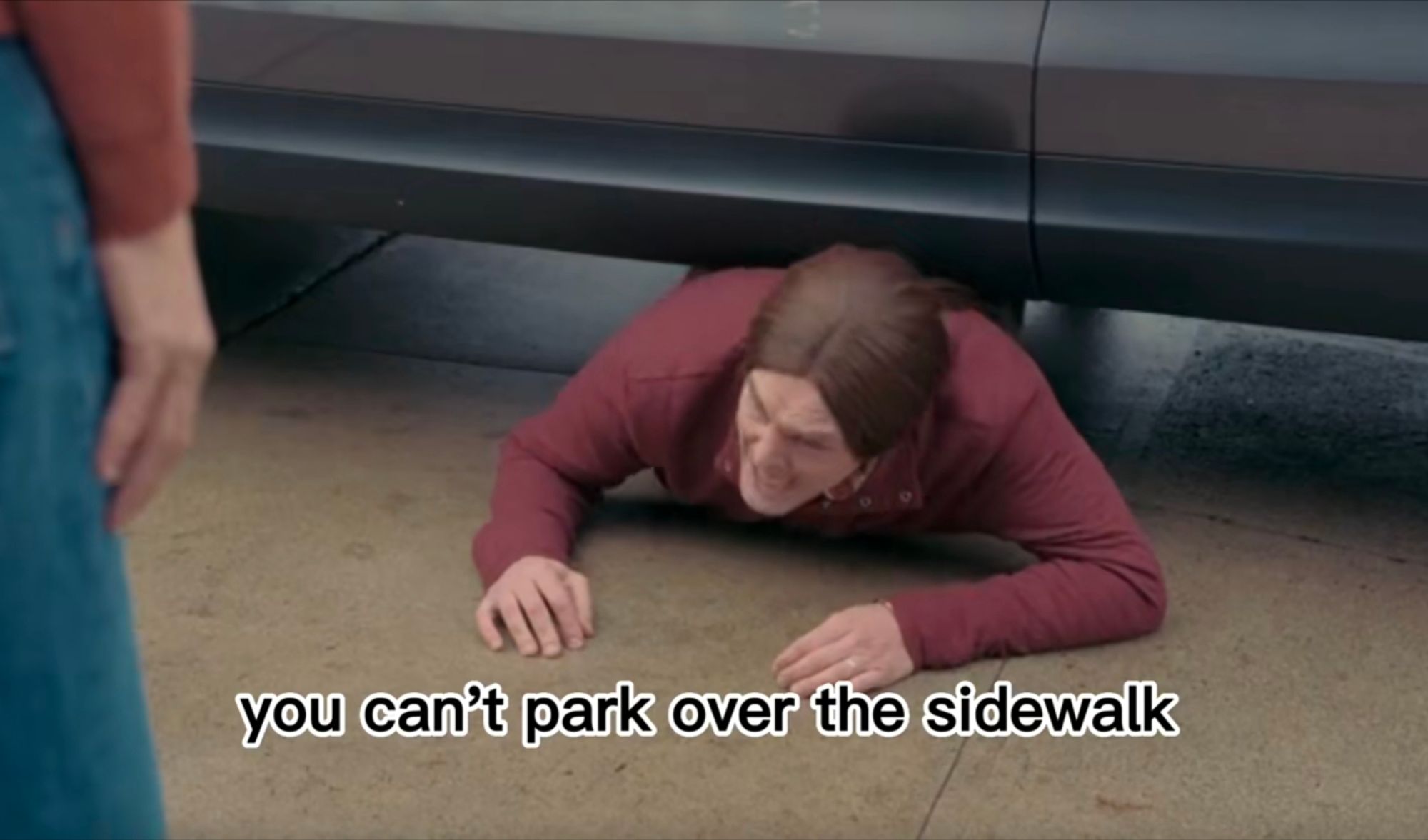 Screen cap from I Think You Should Leave sketch where a long-haired character played by Will Forte has encountered a car parked over the sidewalk and attempted to crawl underneath it to continue his walk. The ponytailed man is pictured under the car yelling "you can't park over the sidewalk!"