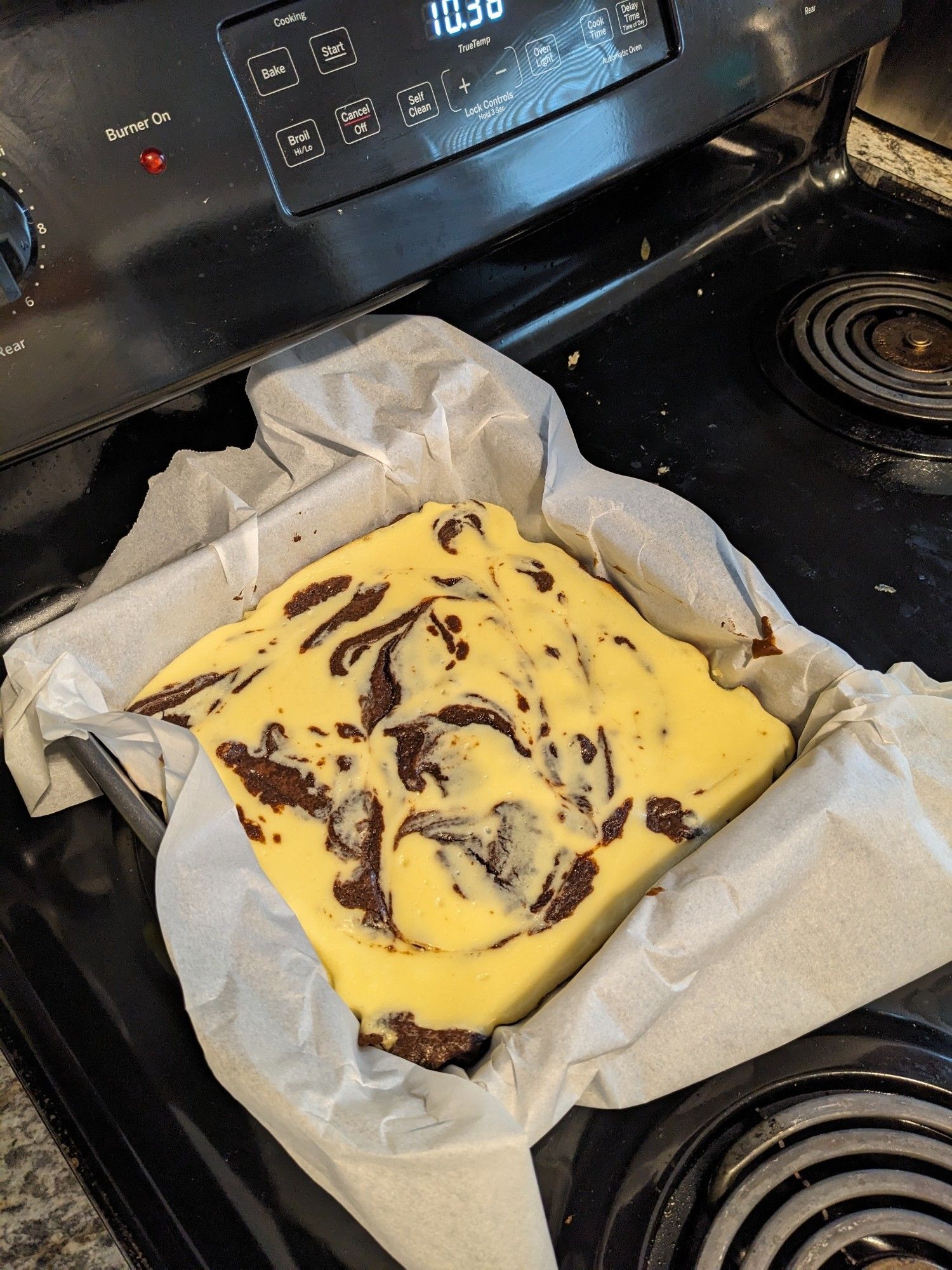 A tray of cheesecake brownies sits cooling on a stove. The off white of the cheesecake is swirled with the chocolate of the brownies below.