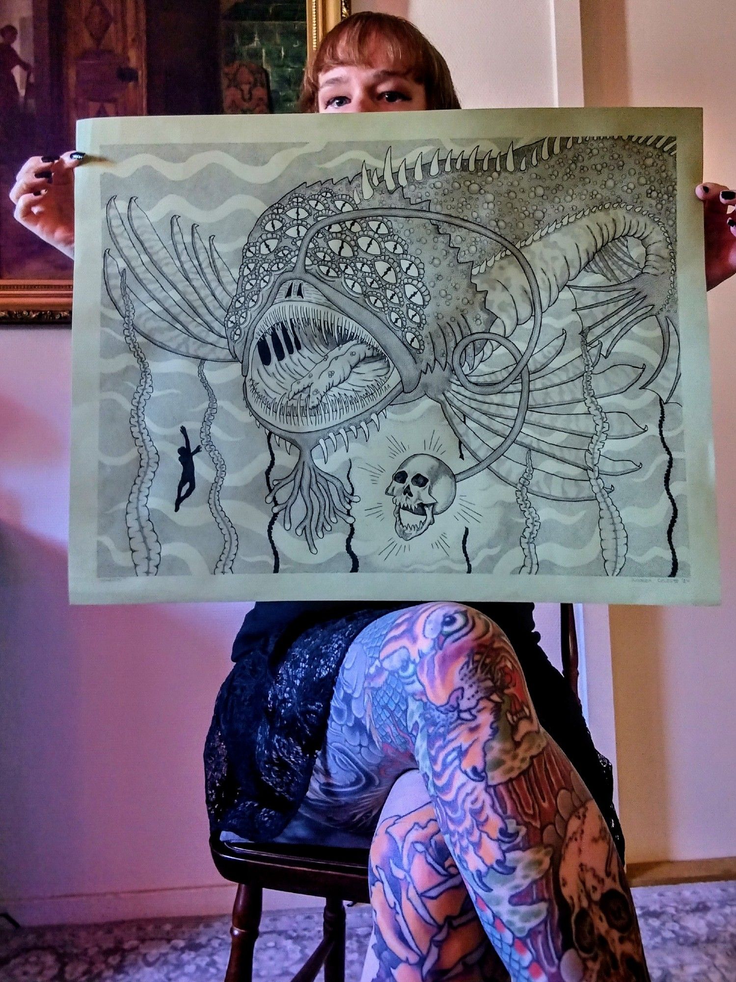 tattooed white woman with red hair and bangs sitting on a wooden chair while wearing a lace skirt and holding up drawing on 70x50 cm green cardstock of marool - a giant anglerfish with many eyes from Scottish folklore. It has lionfish fins and a skull lure. In the bottom left is the silhouette of a drowning person