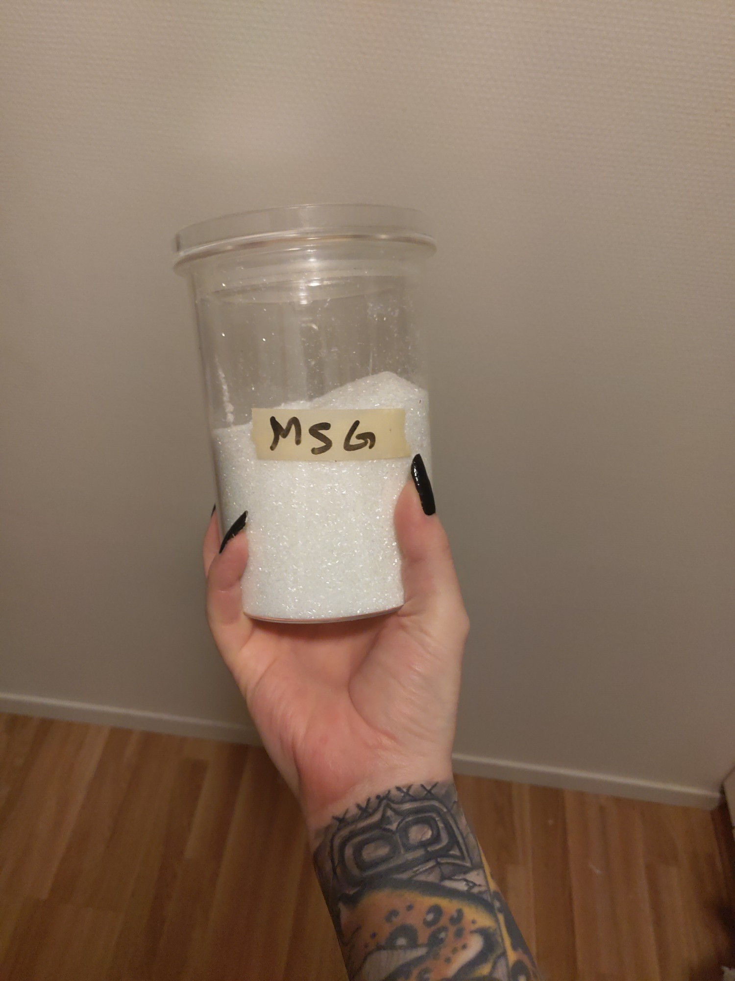 A woman with black nail polish and tattooed wrists holding a big tub of MSG