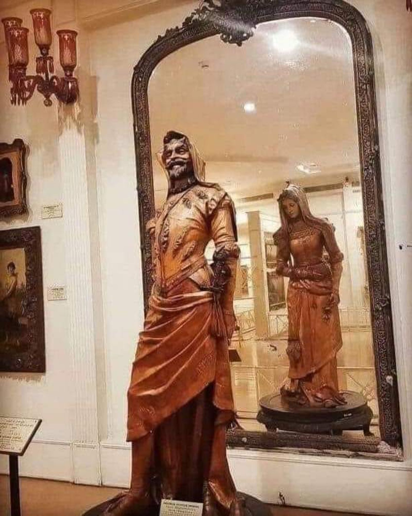 Wooden statue of a roguish male figure on one side and a demure female figure on the other. A large mirror placed behind the statue allows you to see both figures simultaneously.