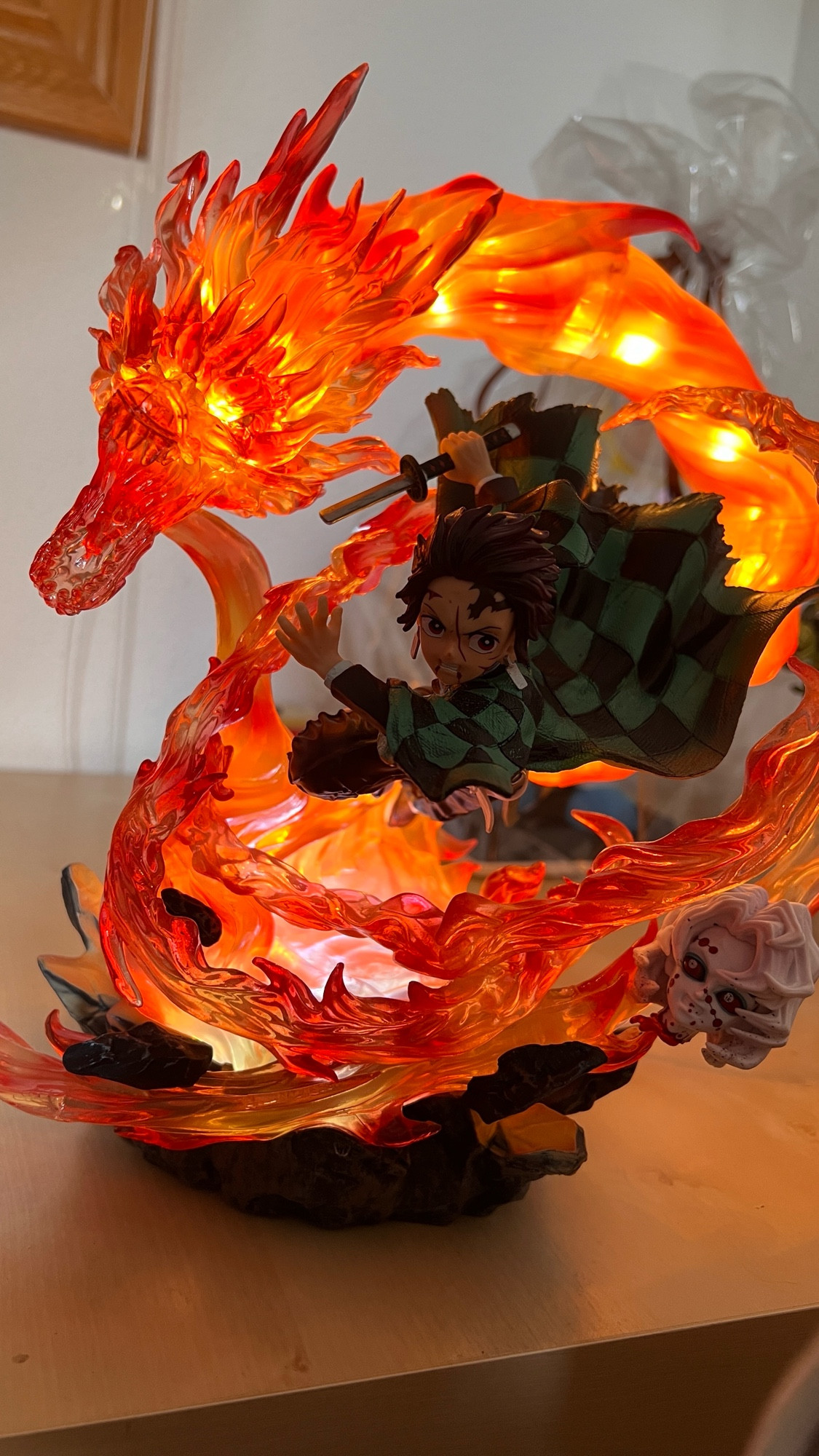 Figura del personaje Tanjiro Kamado, del anime Kimetsu no Yaiba. Se le ve con heridas en la cara, una katana con la hoja rota en su mano derecha y un gran remolino de fuego envolviéndolo a su alrededor, que se encuentra iluminado por luces en su interior. Esta llama termina en la forma de una cabeza de dragón hecho con el propio fuego, junto a la espada de Tanjiro. En un lateral del fuego se observa la cabeza cortada de uno de los demonios de la serie: Rui, que ha sido alcanzado por el ataque de Tanjiro.