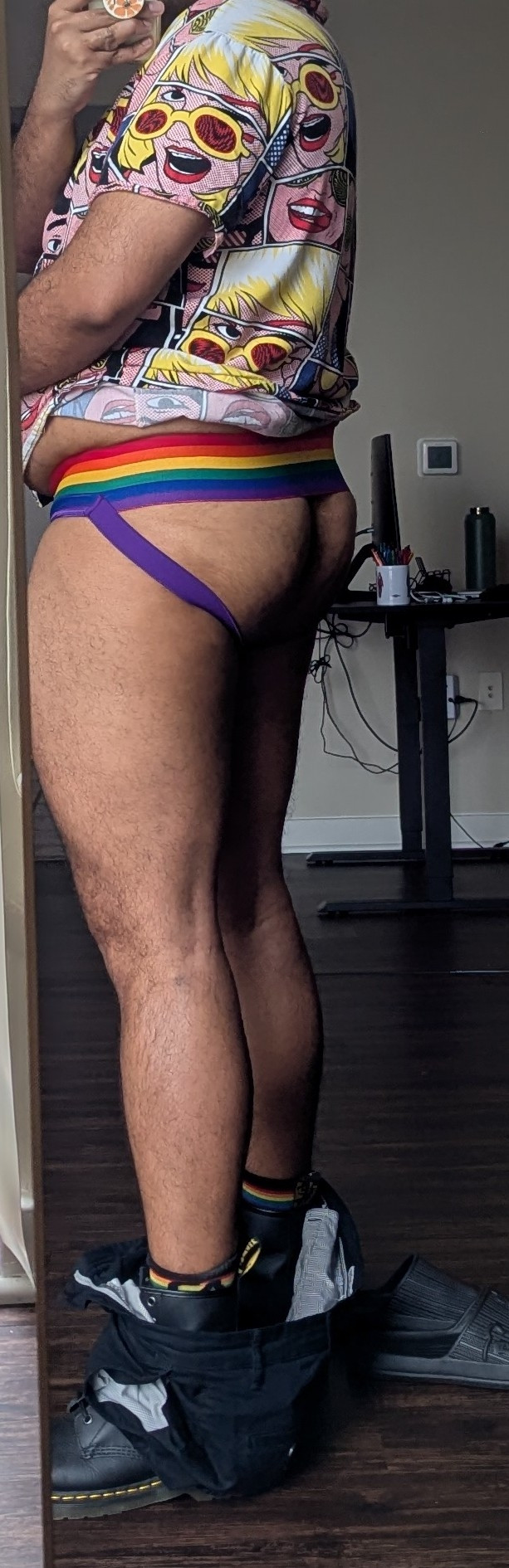 user biguyreacts in the same outfit described above, but with his body slightly turned, shorts around his ankles, revealing his rainbow jockstrap framing his butt. A little belly is poking out as he holds the shirt up so the rainbow band of the jock is visible. 