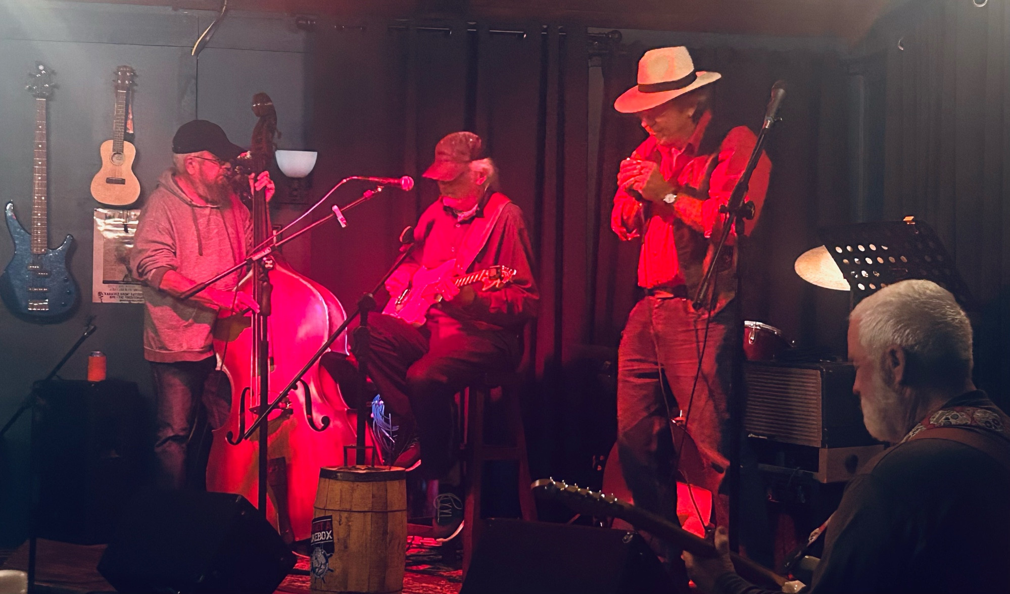 Four men are blues jamming on a small stage in a band shed. 

Three are standing under a red light, the four is seated in the foreground. 