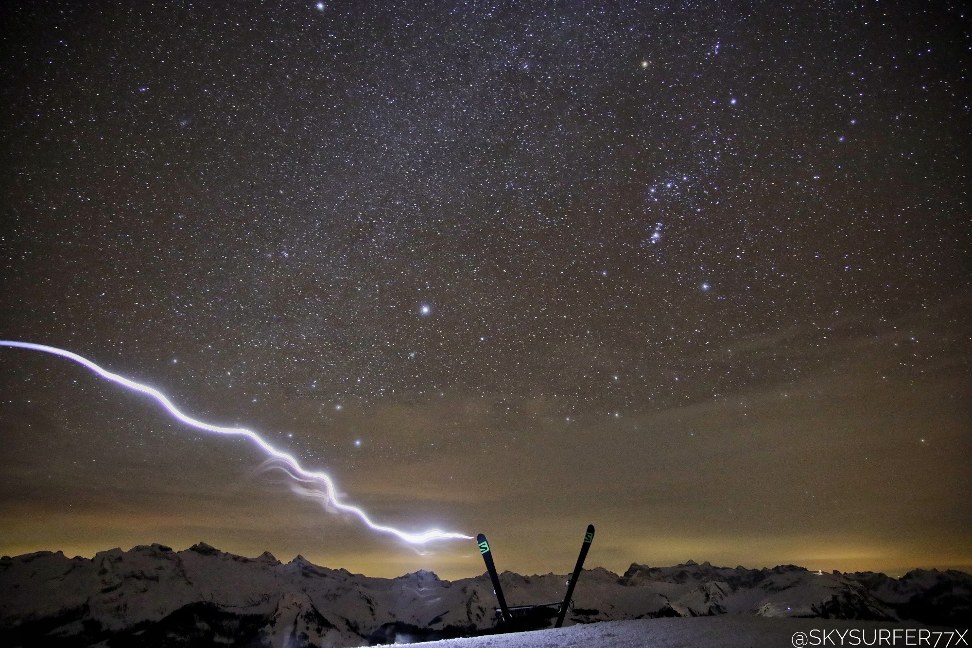 Skis pushed into the snow on a mountain top, to get a chair by using the sticks. While leaving the seat for hot coffee, the headlights accidentally created a streak of light, looking like a lightning strike hitting the skis. In the background a mountain range and the nightsky