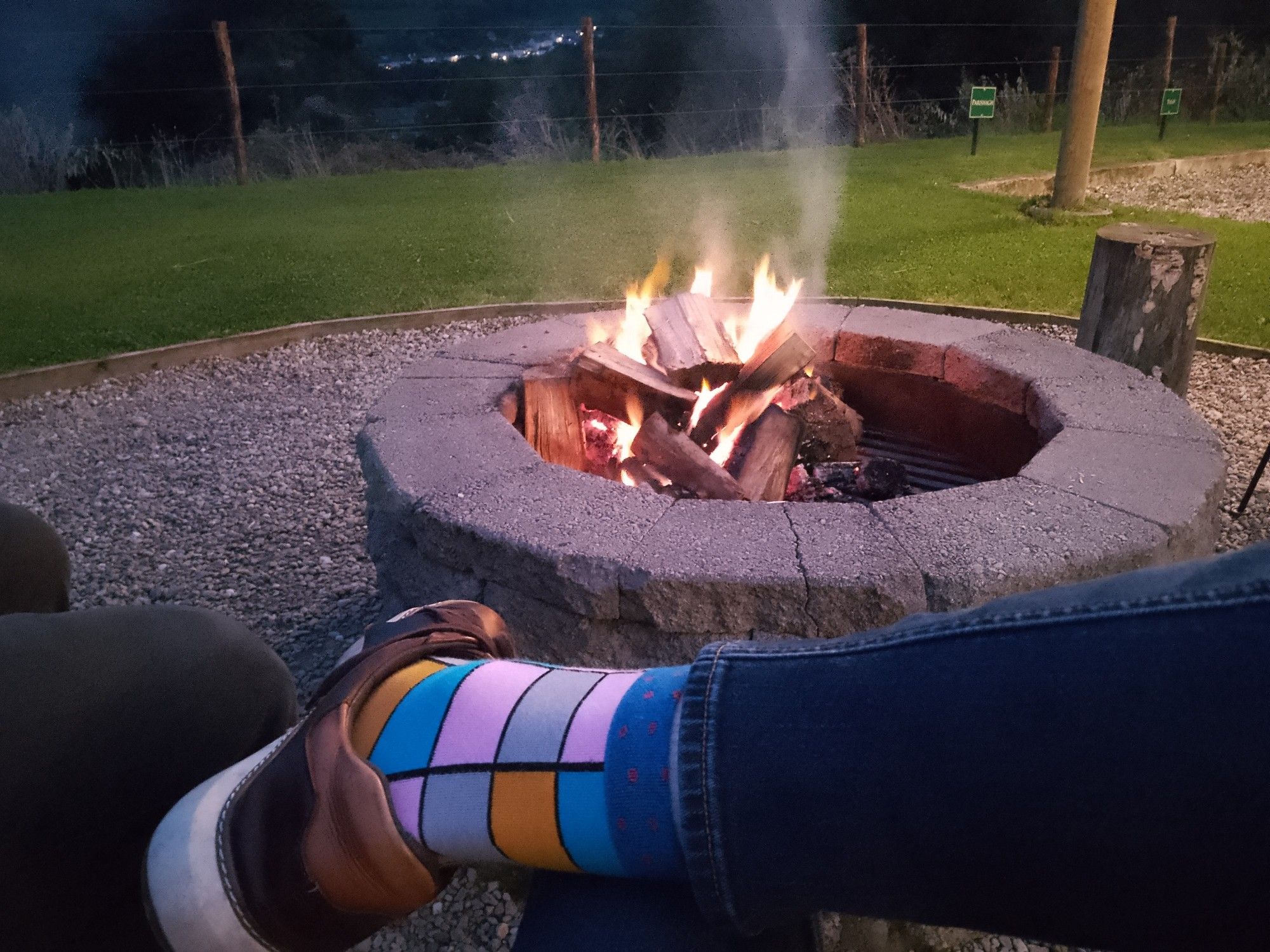 POV of me sitting by the fire pit with my boyfriend outside our pod, watching the flames and smoke rise up as the night draws in