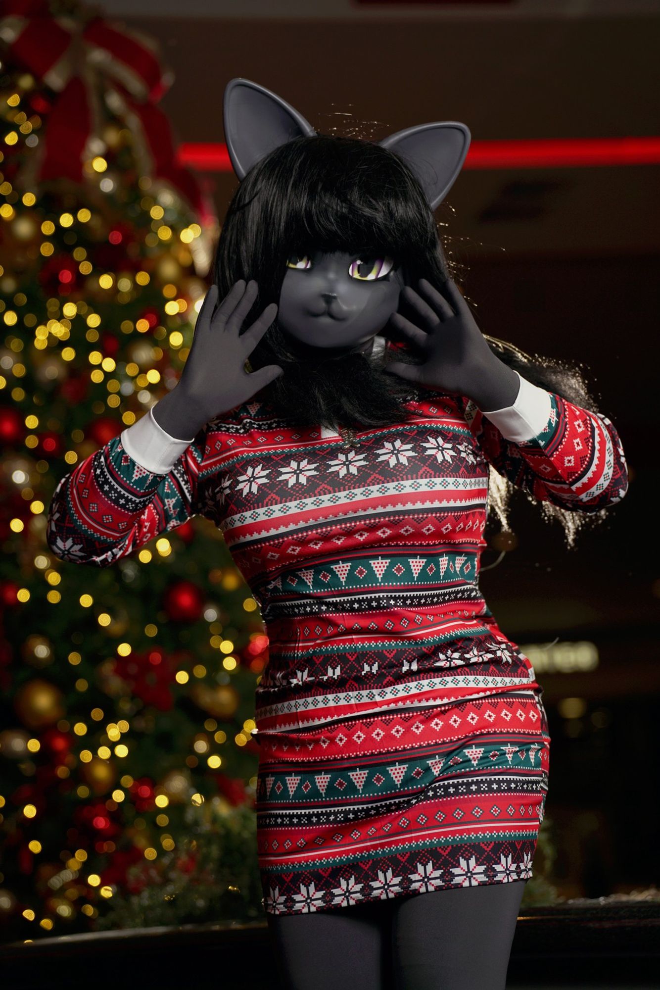 Neko-Nico an anthropomorphic anime-style Kigurumi cat cosplayer is posing with her hands spread open next to her face wearing a short festive Christmas dress in front of a Christmas tree.