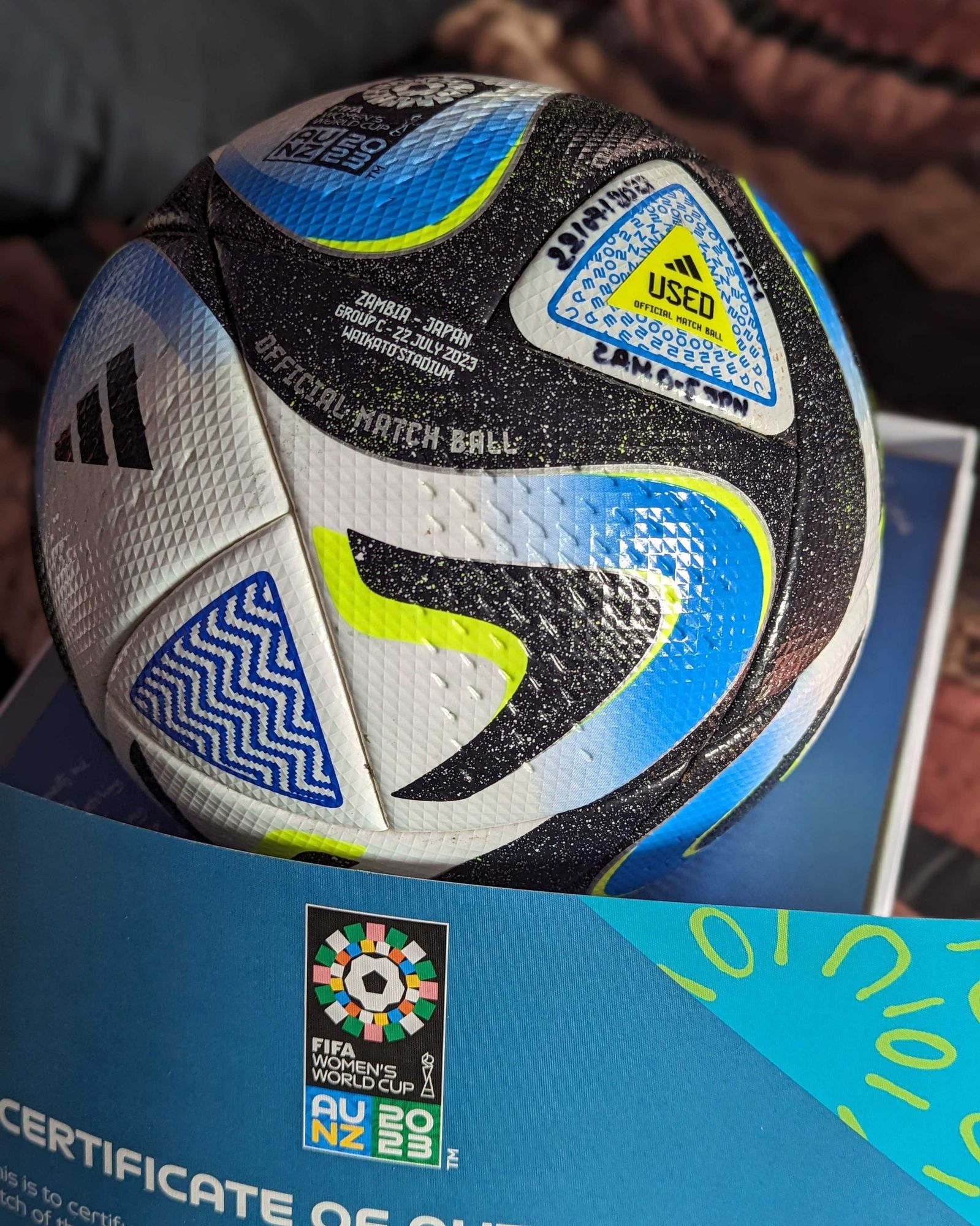 A white, blue, and shimmery black swirled Adidas Oceaunz soccer ball with neon yellow accents, nestled on a blue and white presentation box.