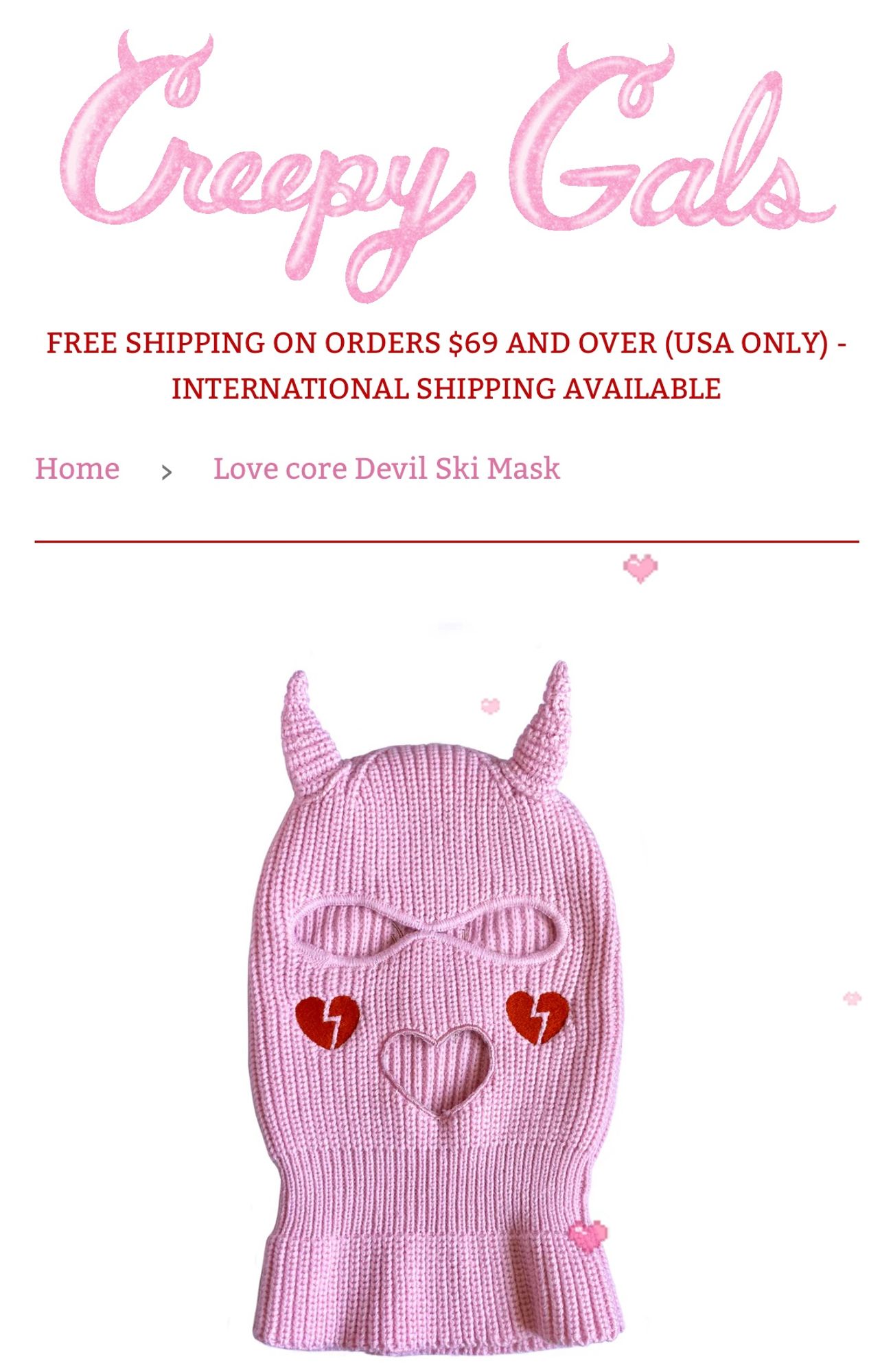 A light pink ski mask with devil horns. It has red embroidered broken hearts on the cheeks and a mouth cut out in the shape of a heart. 