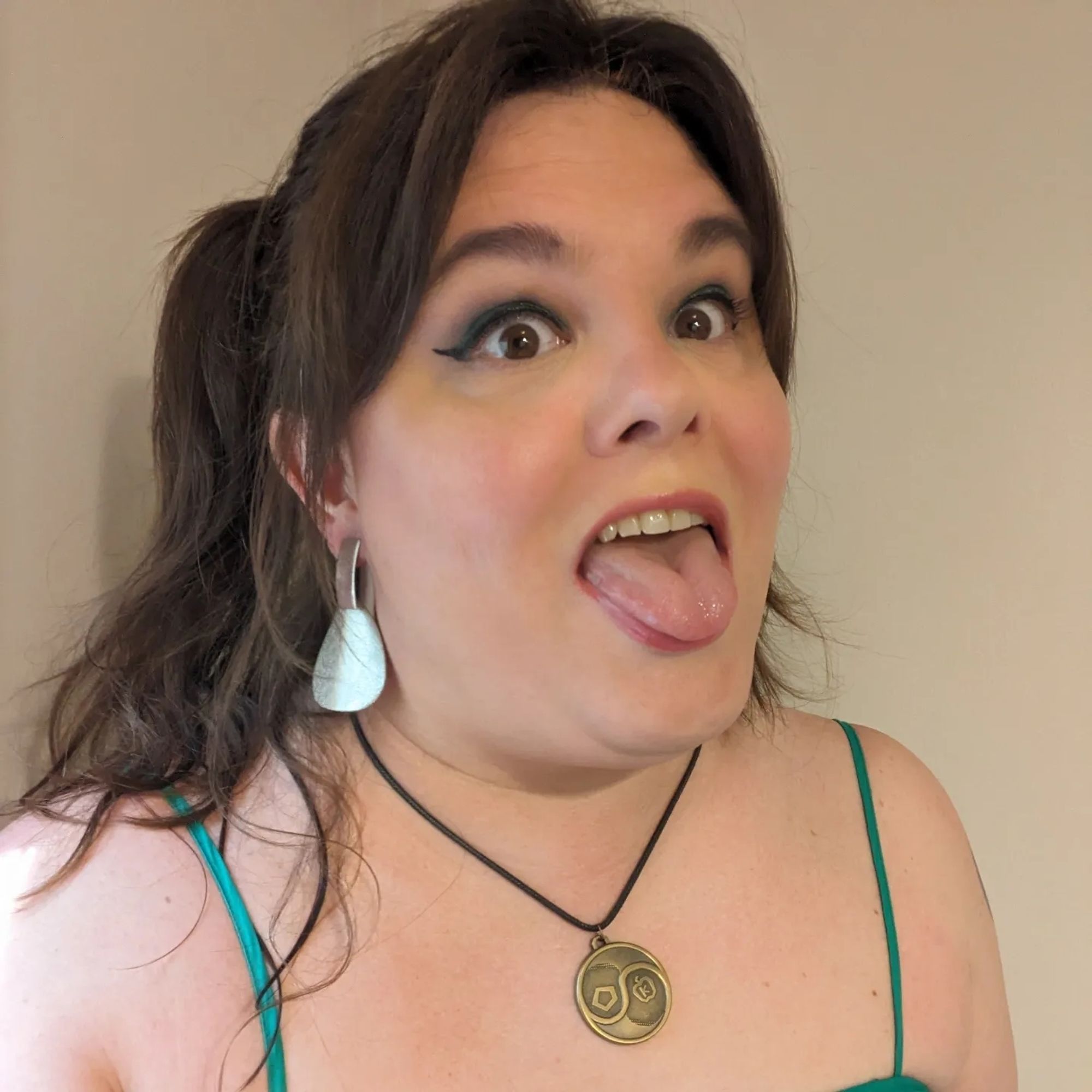 Lydia from the chest up in a low-cut green dress. her hair is in messy pigtails and she is wearing large dangly earrings that look like silver fishing lure spinners. she has heavy eye makeup on as well as blush and pink lipgloss; she is sticking her tongue out of her open mouth, her eyes widely staring up and to the right, off camera. she wears around her neck a brass Sacred Chao medallion