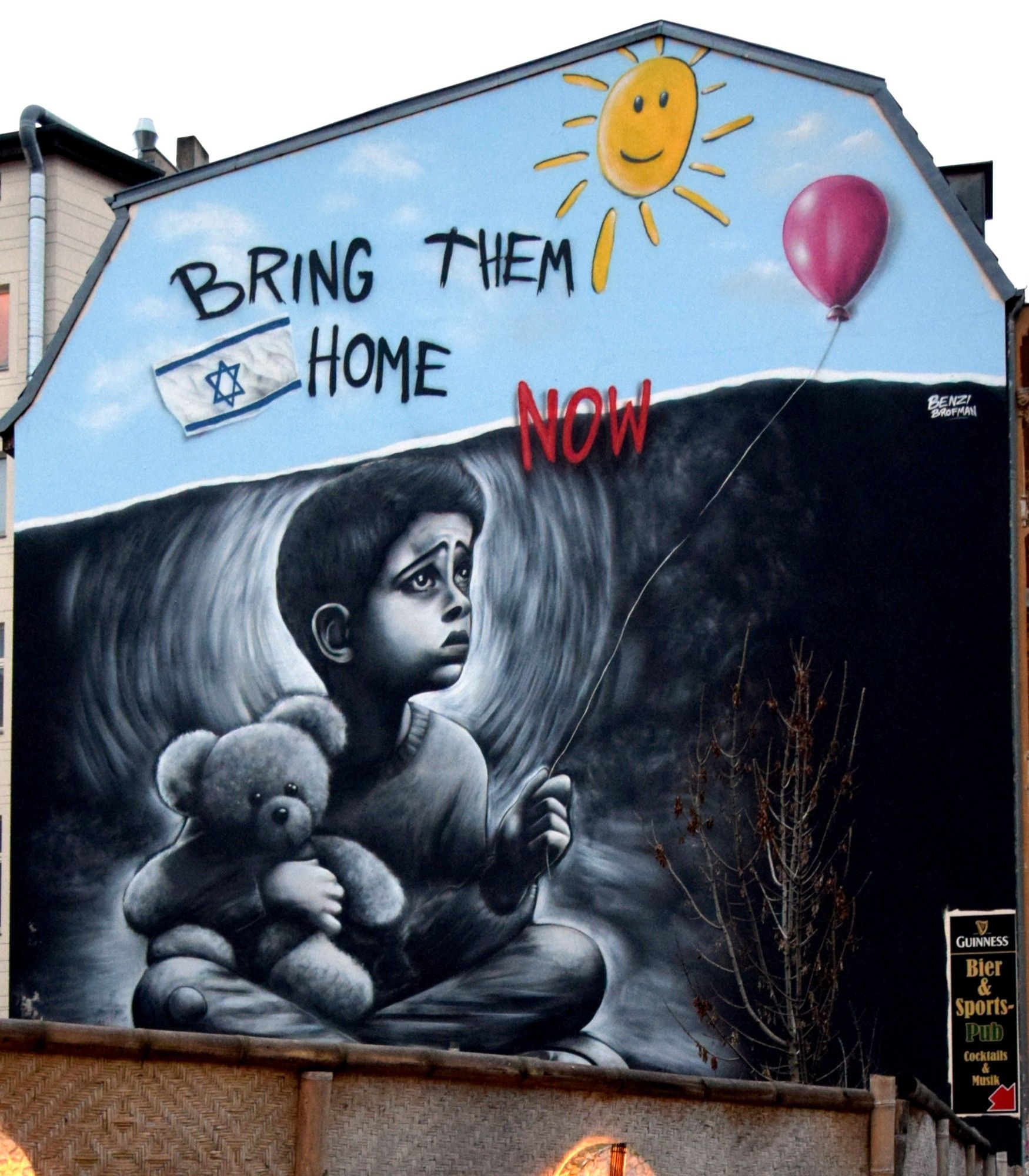 Graffiti an einem Haus: ein Kind mit Teddybär und Luftballon in einem dunklen Tunnel. Der Ballon ragt in einen blauen Himmel.
Daneben der Slogan "Bring them home now" und eine Israelfahne.