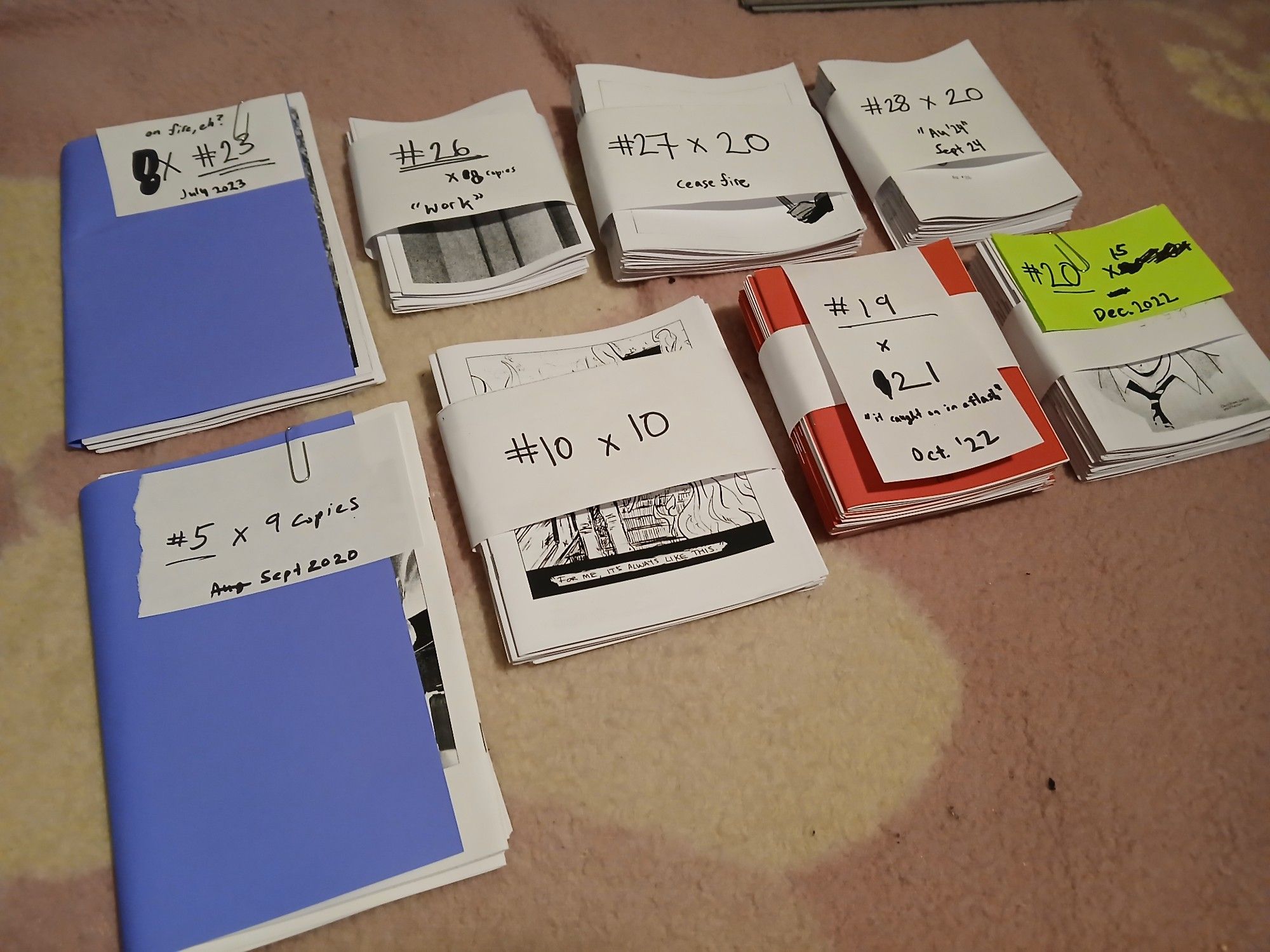 Eight stacks of zines with makeshift paper rings and inventory labels. Blue, red, green.