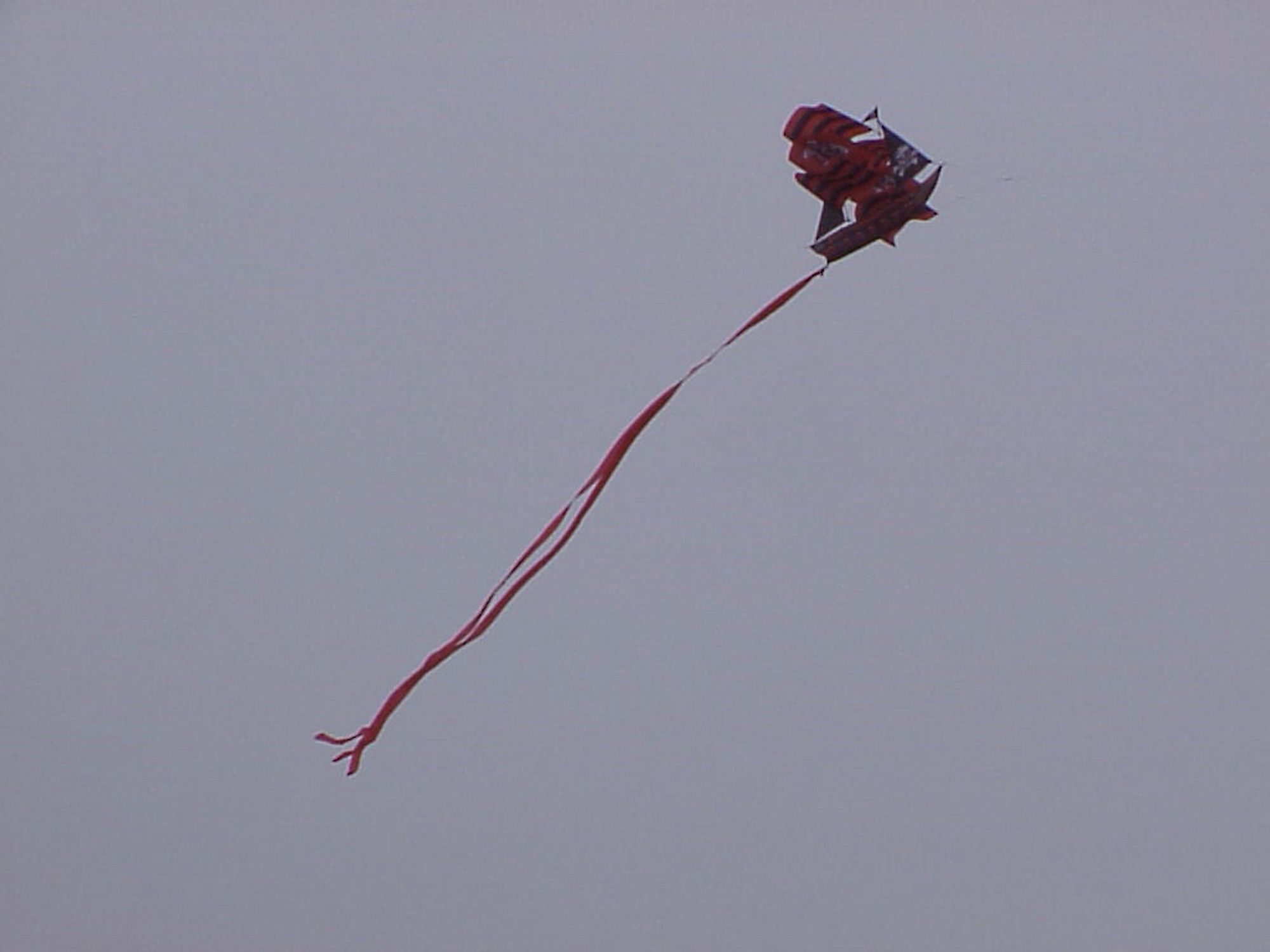 A kite that looks like a pirate ship, in the sky. The pirate ship is red and black, and it has multiple flags and masts. It has two tails attached at the back of the ship.