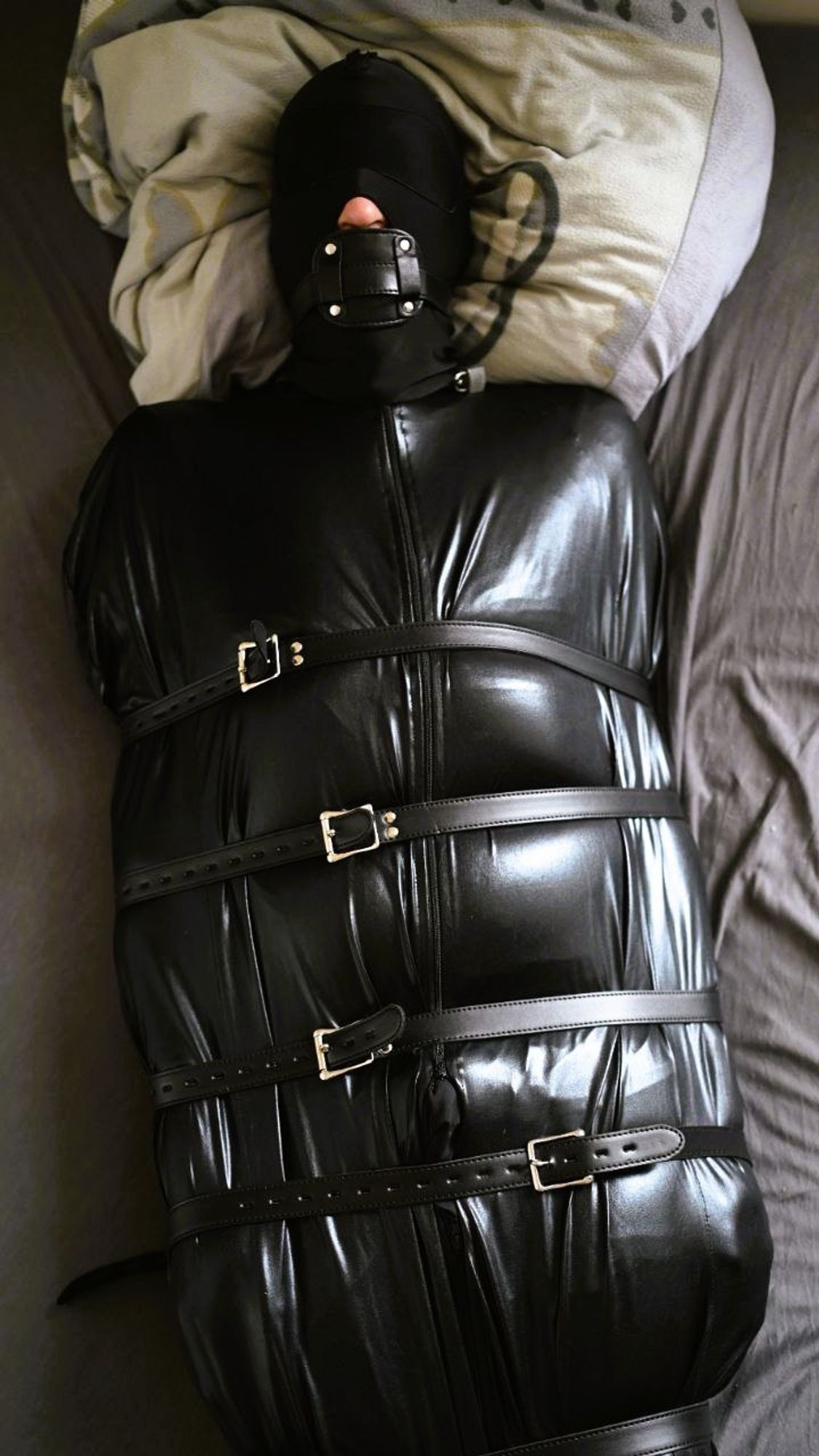 Detail of a person in a black bondage sleepsack on a mattress, with leather straps closed around it, wearing a hood and gagged.