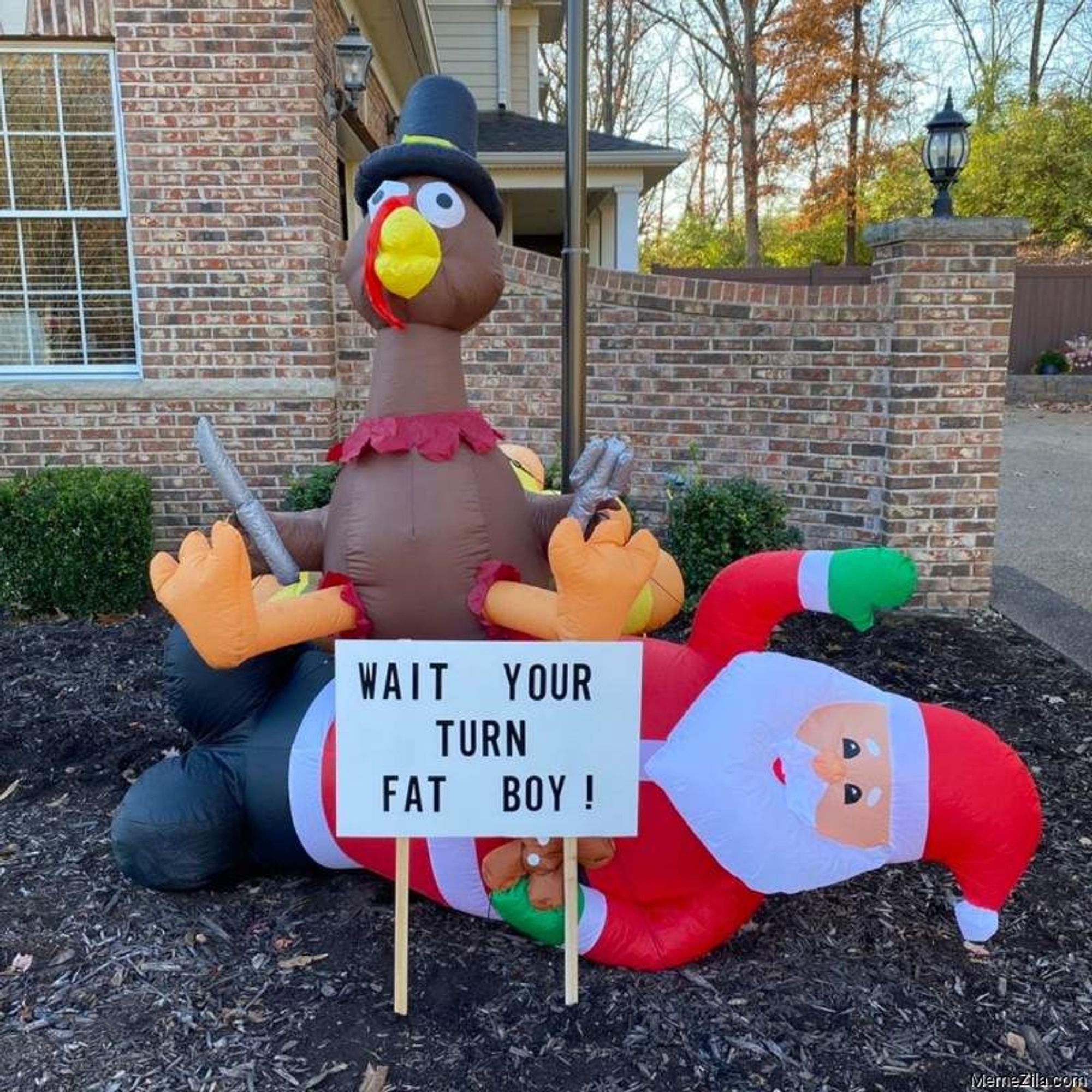 An inflatable turkey lawn ornament sitting on an inflatable Santa lawn ornament with a sign in front that says "Wait your turn, fat boy!"