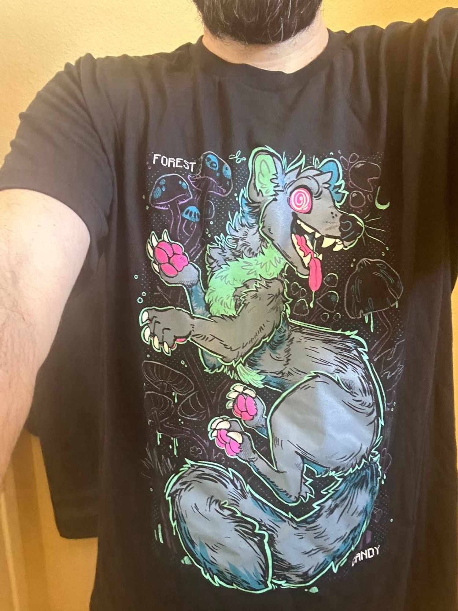 Me wearing a black shirt with some sort of weasel/marten critter (he’s a gray-blue with a pastel green underbelly and pink pays, tongue and swirly eyes) that’s high on “Forest Candy” as the shirt says- which are implied to be psilocybin mushrooms.