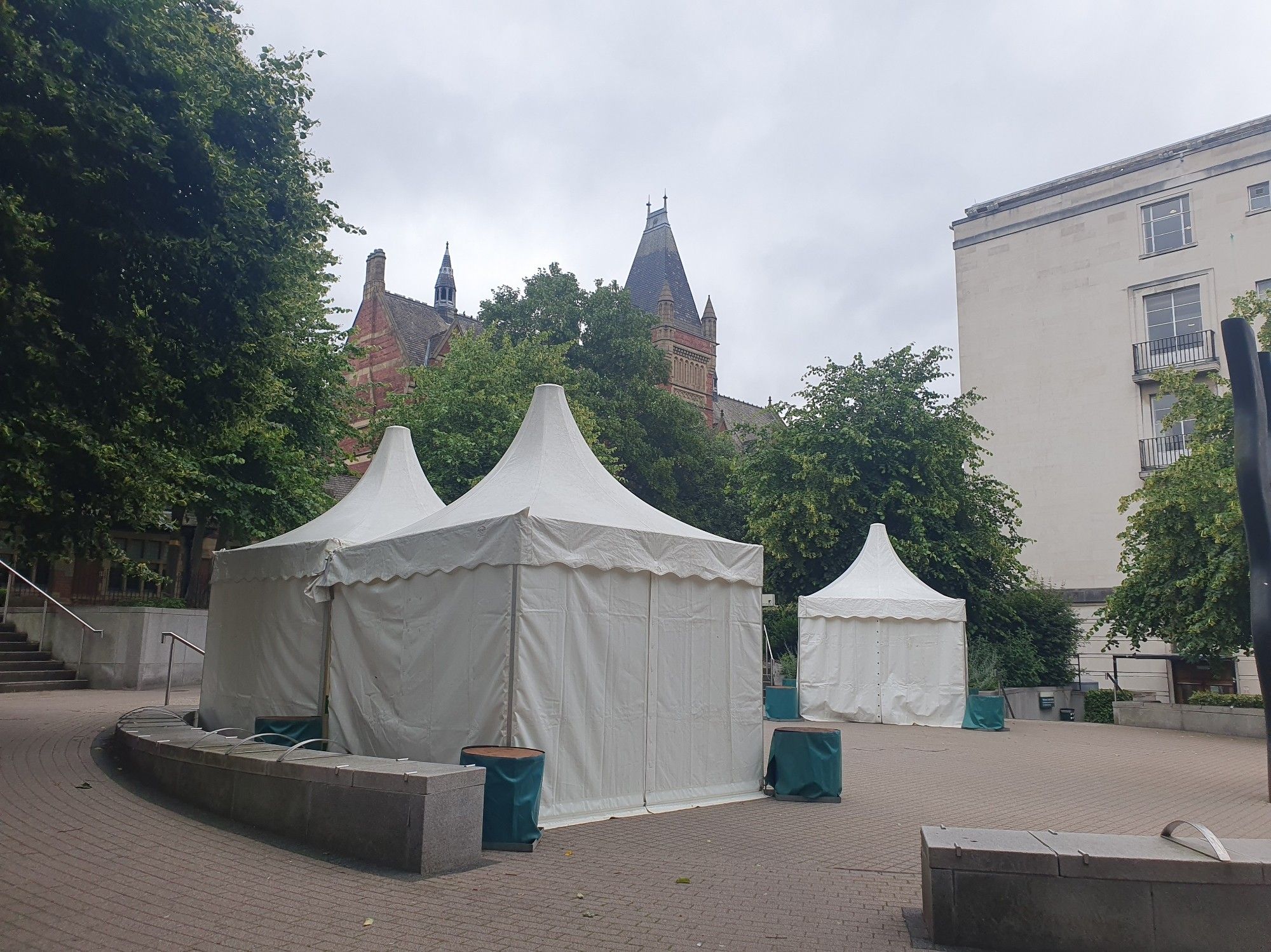 Photo shows three white, square tents with pointed roofs, in a courtyard surrounded by trees