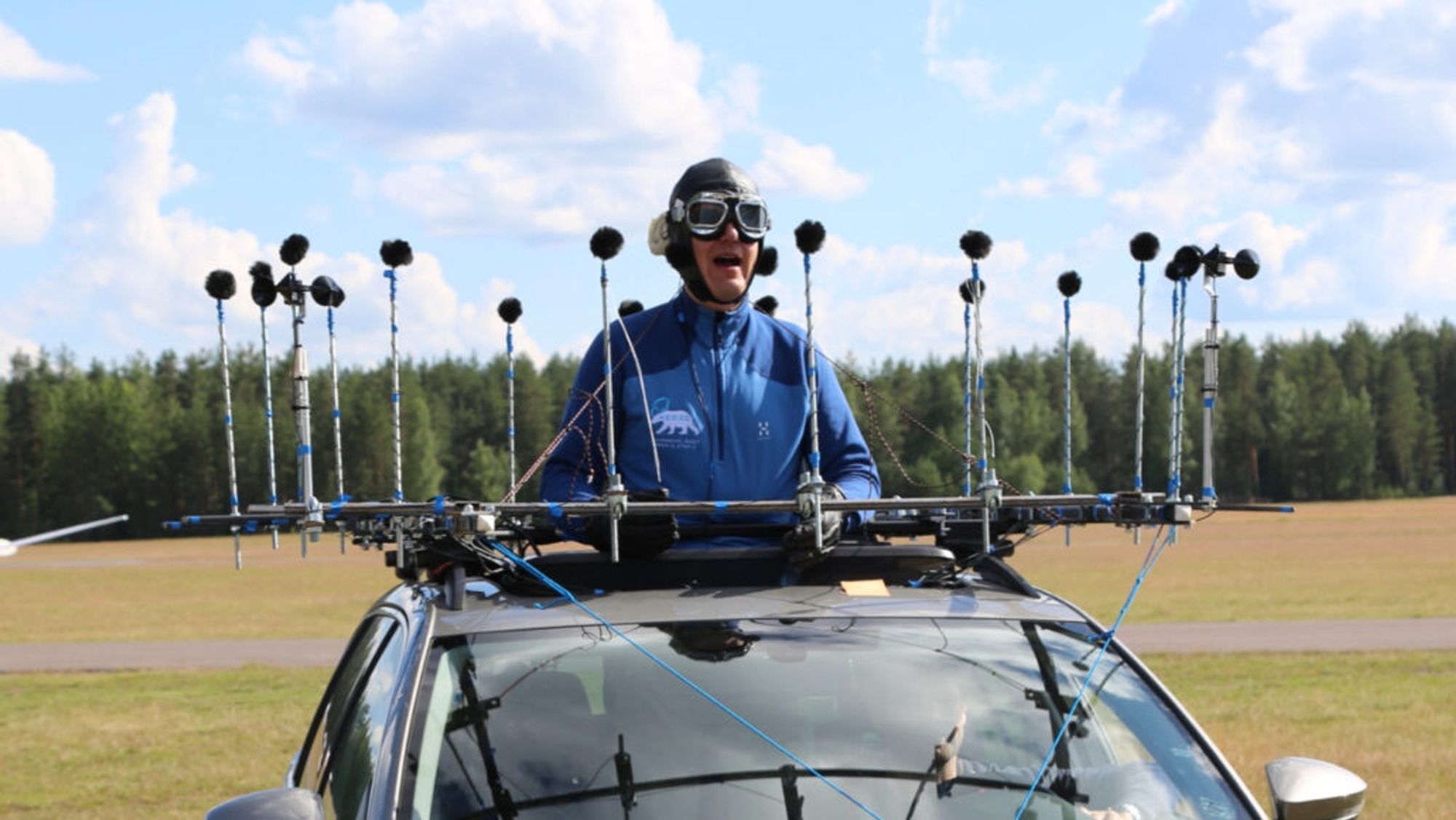 A photo of Ville Pulkki, an older white man, wearing a flight helmet, goggles and a blue jacket . His upper body is seen through the sunroof of a car. There is a ring of several small microphones surrounding him.