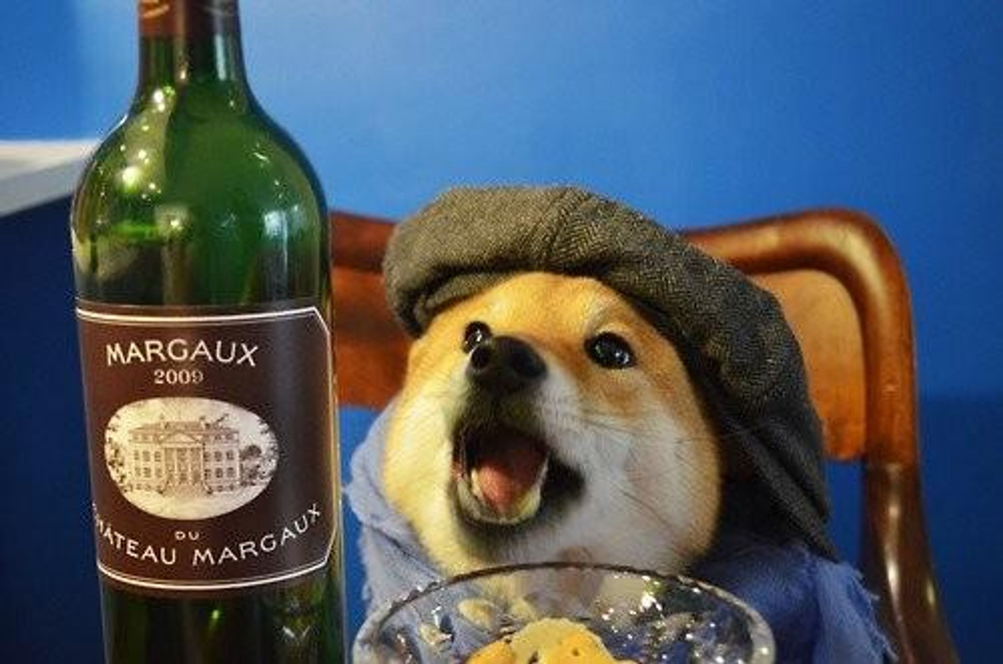 A dog wearing a flat cap is sitting in a chair with a glass of wine in front of him. A bottle of wine is in the background. The dog is looking at the camera with its mouth open and its tongue sticking out. The image is a humorous take on the classic image of a man sitting in a chair with a glass of wine.