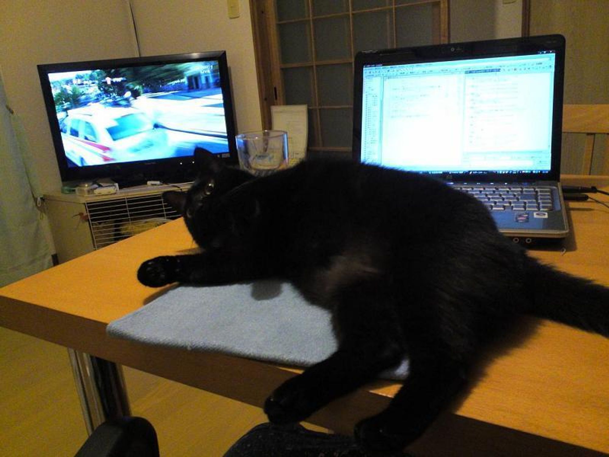 A black cat lies sprawled out on a table in front of a laptop computer and a TV, clearly in charge of the home office.