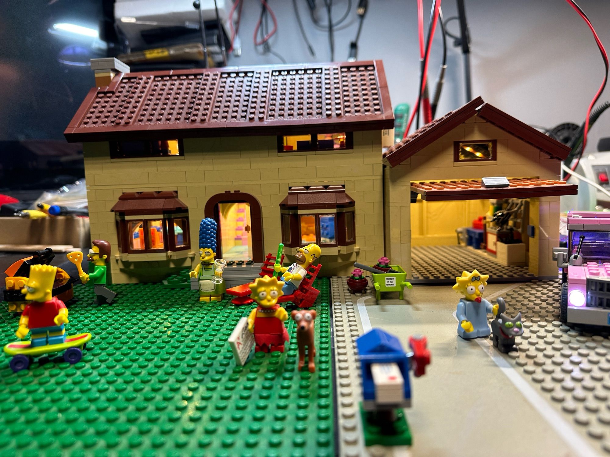 A Lego model of the Simpsons house with the Simpsons out in front and the car parked next to the open garage. All lit up with LED lighting.