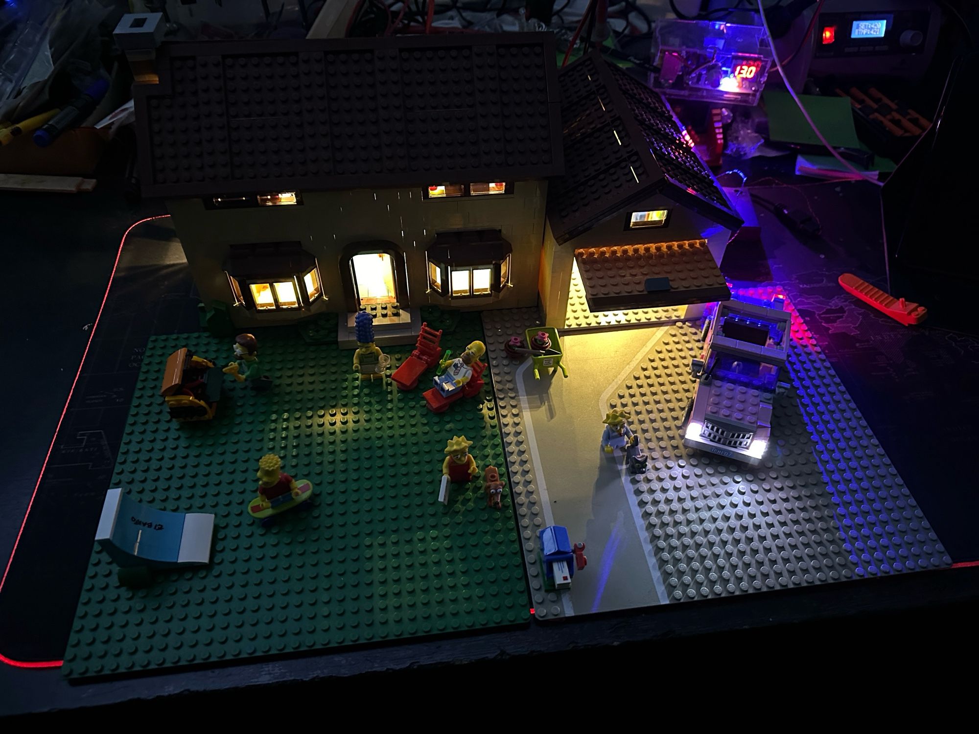 A Lego model of the Simpsons house with the Simpsons out in front and the car parked next to the open garage. All lit up with LED lighting.