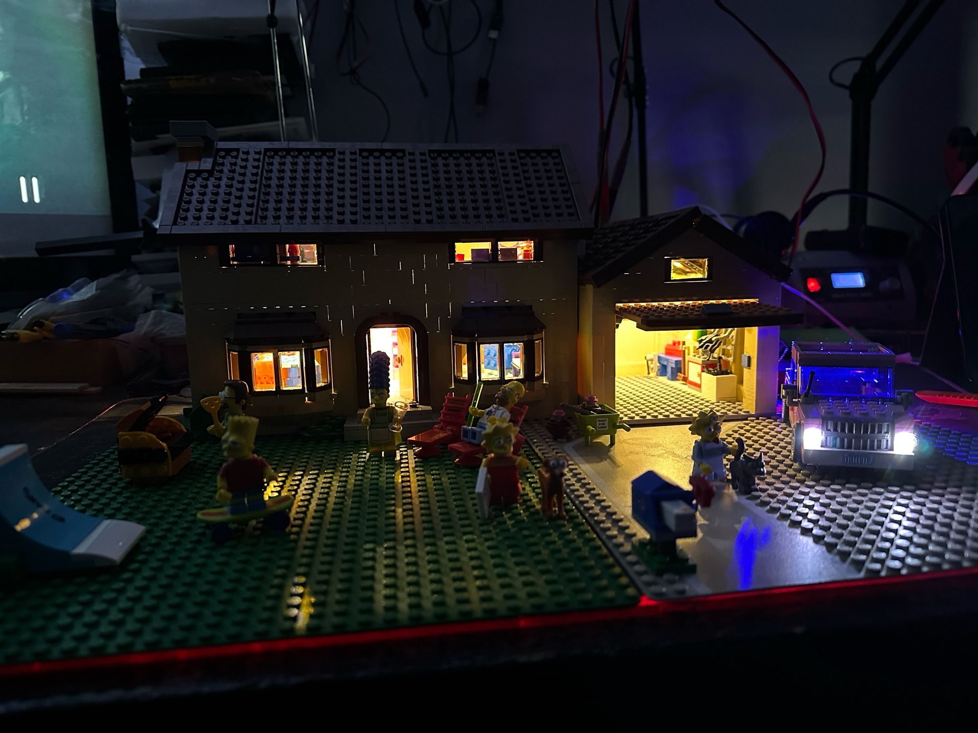 A Lego model of the Simpsons house with the Simpsons out in front and the car parked next to the open garage. All lit up with LED lighting.