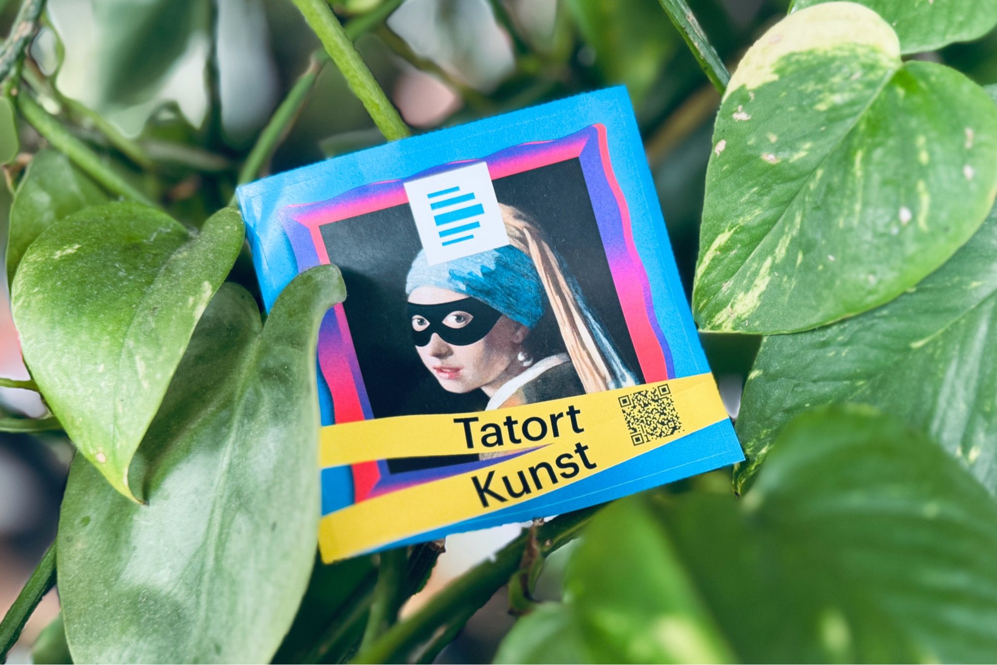 Ein Aufkleber des Podcasts Tatort Kunst zwischen grünen Blättern.