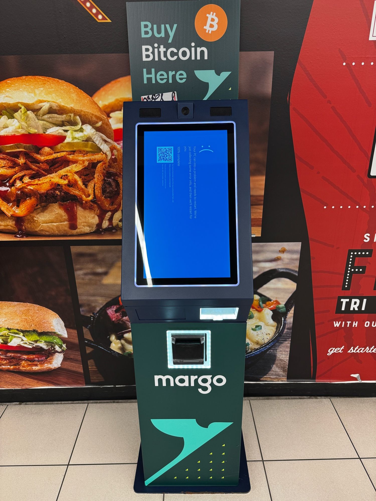 A Mango-brand bitcoin ATM in a mall. It has a large sign at the top that says “Buy Bitcoin Here”. The screen doesn’t show user interface, instead it is displaying a Windows Blue Screen of Death (BSOD) with the :( emoticon prominently displayed).