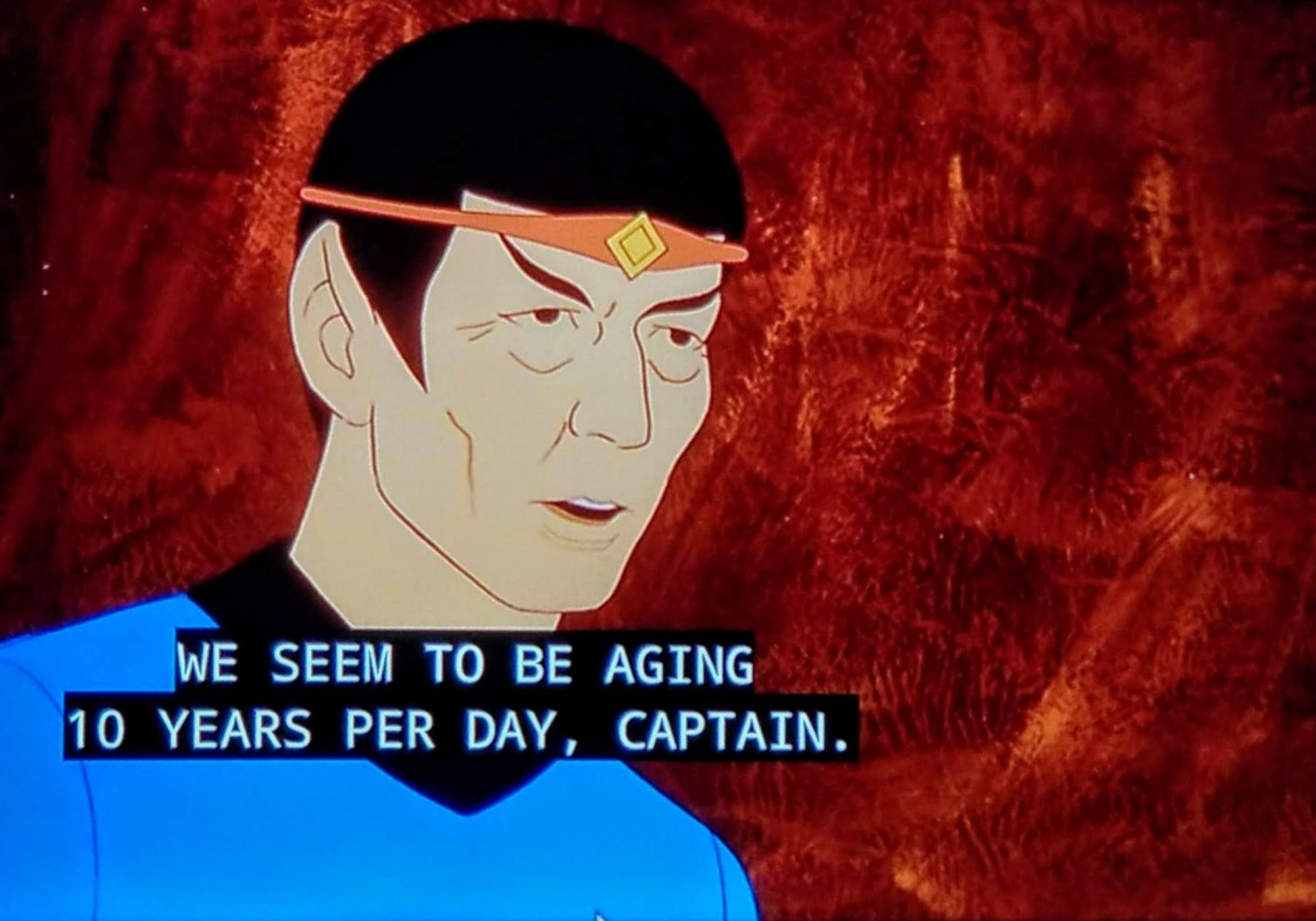 Star Trek The Animated Series scene. (From the olden days, not Lower Decks) Here we see Spock, but he's very old! He's in his normal blue uniform and is also wearing a headband with a diamond shaped golden stone or something right over this forehead. Kind of a hippie looking item. Besides being old the other noteworthy thing here is he's standing against a red wall that looks like somebody took construction paper and a red marker and just scribbled all over it for about 2 hours. Closed captioned reads, "We seem to be aging 10 years per day, Captain."