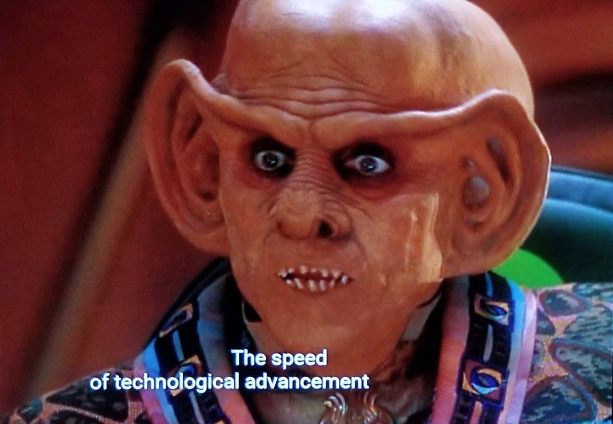 DS9 scene. We're in Quark's bar I assume, and we're close in on Quark. He's a ferengi alien barkeep who wears fancy jackets with patterns and colours that look like hotel carpet. He has dark, sunken eyes and is bald with a big bulbous head. Also has giant ears. But the ears also form a line across his forehead and meet between the eyes so it's like a unibrow but with giant ears. He also has sharp teeth. Ok, enough about Quark. Here, he's speaking with some energy. Closed caption reads, "The speed of technological advancement isn't nearly as important as short term quarterly gains."