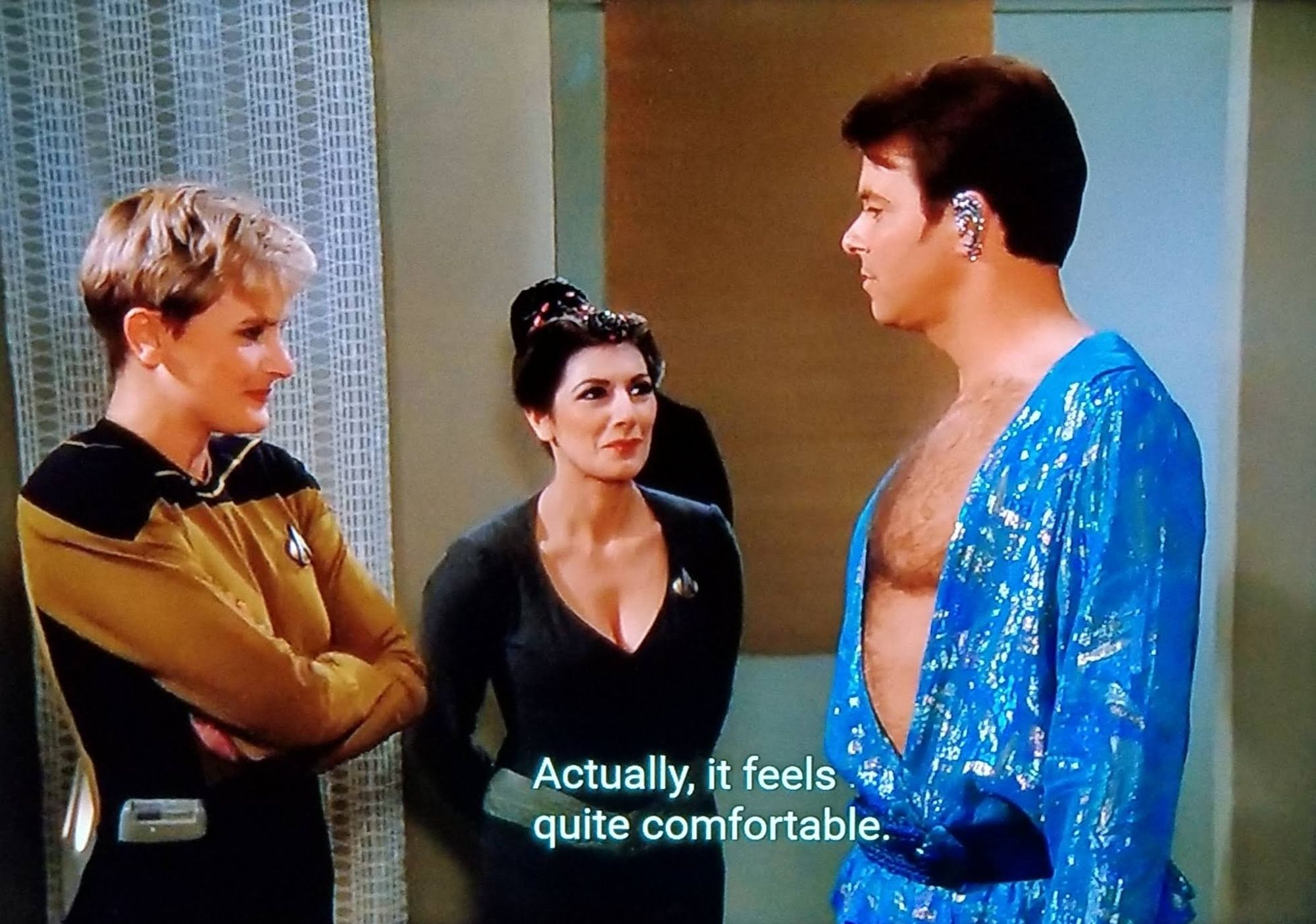 TNG scene. Looks like we're on an away mission somewhere in a room that is pretty unremarkable. We see Will Riker, Deanna Troi, and Tasha Yar. Troi is in her pre-laterseason-actual uniform leotard counseling outfit and Yarrr is in gold normal uniform. They're both stifling giggles and holding back smiles as they look on at Riker, who is standing and facing them. Riker is wearing a long shimmery glittery silky lil' number with a wide open chest plunge down to the waist line displaying his hairy chestal region proudly. He's also wearing a bedazzled earring. Closed caption reads, "Actually, it feels quite comfortable."