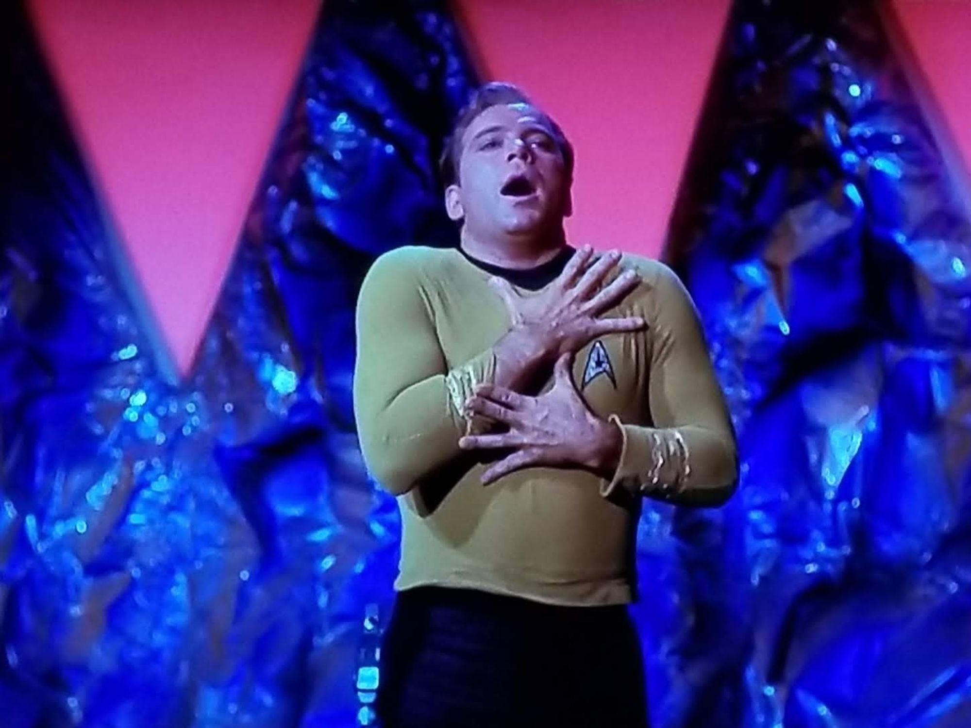 TOS scene. We're in some kind of cave but the rocky wall is blue and there's upside down pink triangles decorating across. Kirk is standing in uniform. He's making an "O face" and clutching himself, FEELING himself. He's holding his chestal region with jazz hands like he's an hour into MDNA trip. No caption.