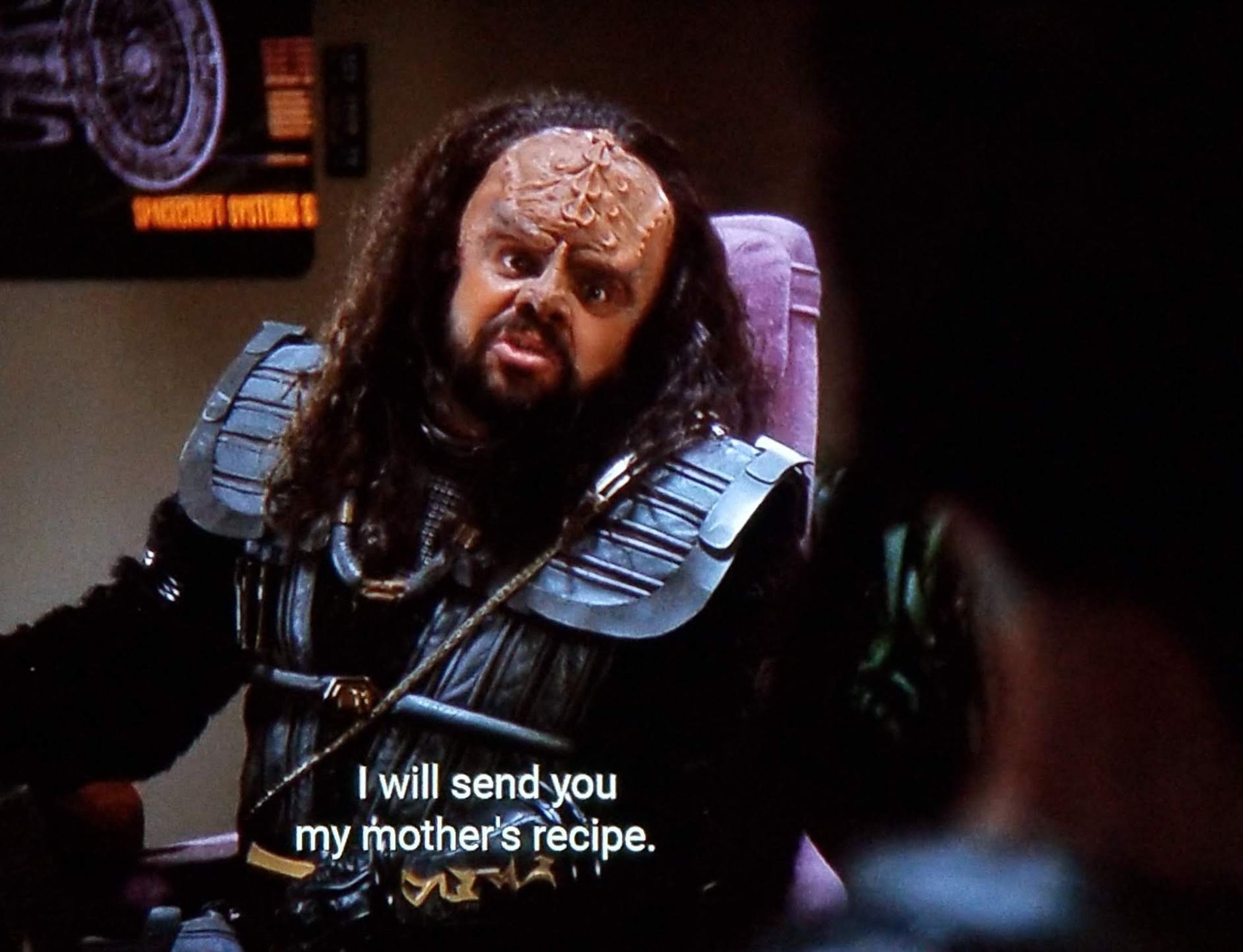 TNG scene. We're in the Enterprise conference room. We see a Klingon in normal silver black uniform that looks a bit like armor. He has sharp ridged loaf along the forehead and long curly hair past the shoulders stemming from an aggressively receding hairline. He's turned from the table with energetic eyeballs and speaking to someone across the room. Closed caption reads, "I will send you my mother's recipe."