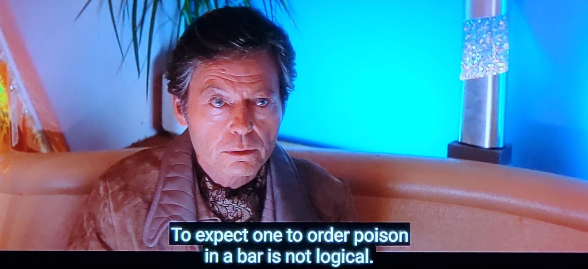 Scene from Star Trek III, The Search For Spock. We're in a futurey bar, looking at Bones sitting in a booth. There's a plant behind him and some crystal ornamental fixtures and mood lighting in a soft blue. The booth itself is sort of a beige/tan, a curious choice in both color. Bones is in a leather jacket with a 70s looking paisley brown silk scarf around his neck, which was the style at the time. He looks slightly bugged out and intense. Closed caption reads, "To expect one to order poison in a bar is not logical."