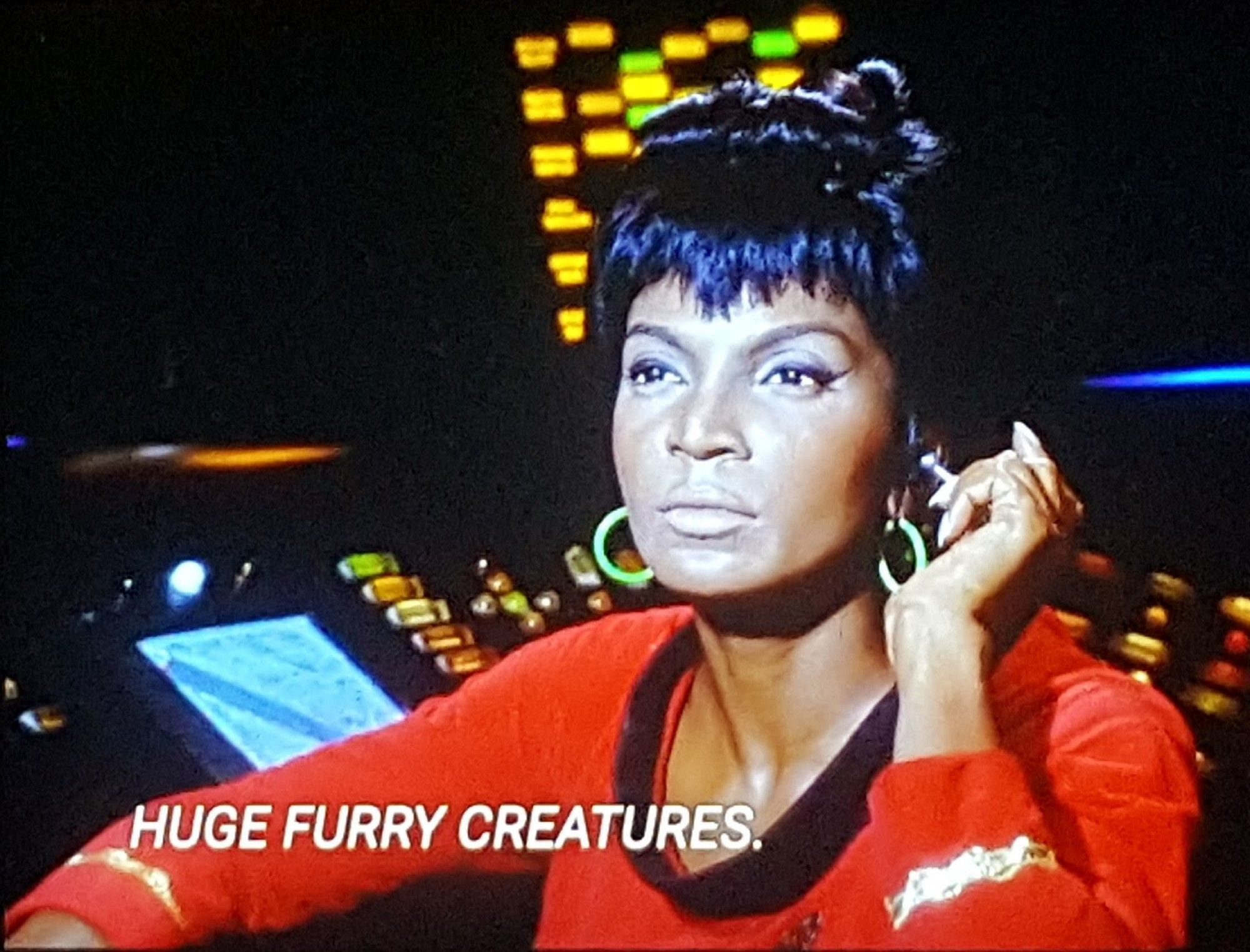 TOS scene. Pictured is Uhura, in red uniform, sitting at her communications station on the bridge. She's turned out from her station and touching her silver Bluetooth ear piece thingy like she's listening to something about huge furry creatures. Closed caption reads, "HUGE FURRY CREATURES."