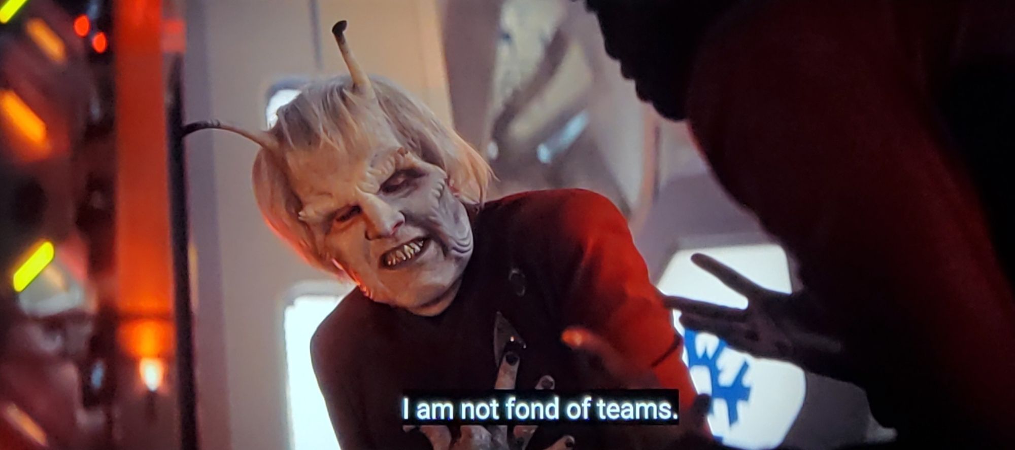 Strange New Worlds scene. Hemmer is shown, in red uniform. He's a alien fella, grayish pale skin, white hair, 2 big antennas popping out of his head. Also has some rippling around his cheeks, palpatine-esqu. Here, he looks kind of pained/doubled over. Closed caption reads, "I am not fond of teams."