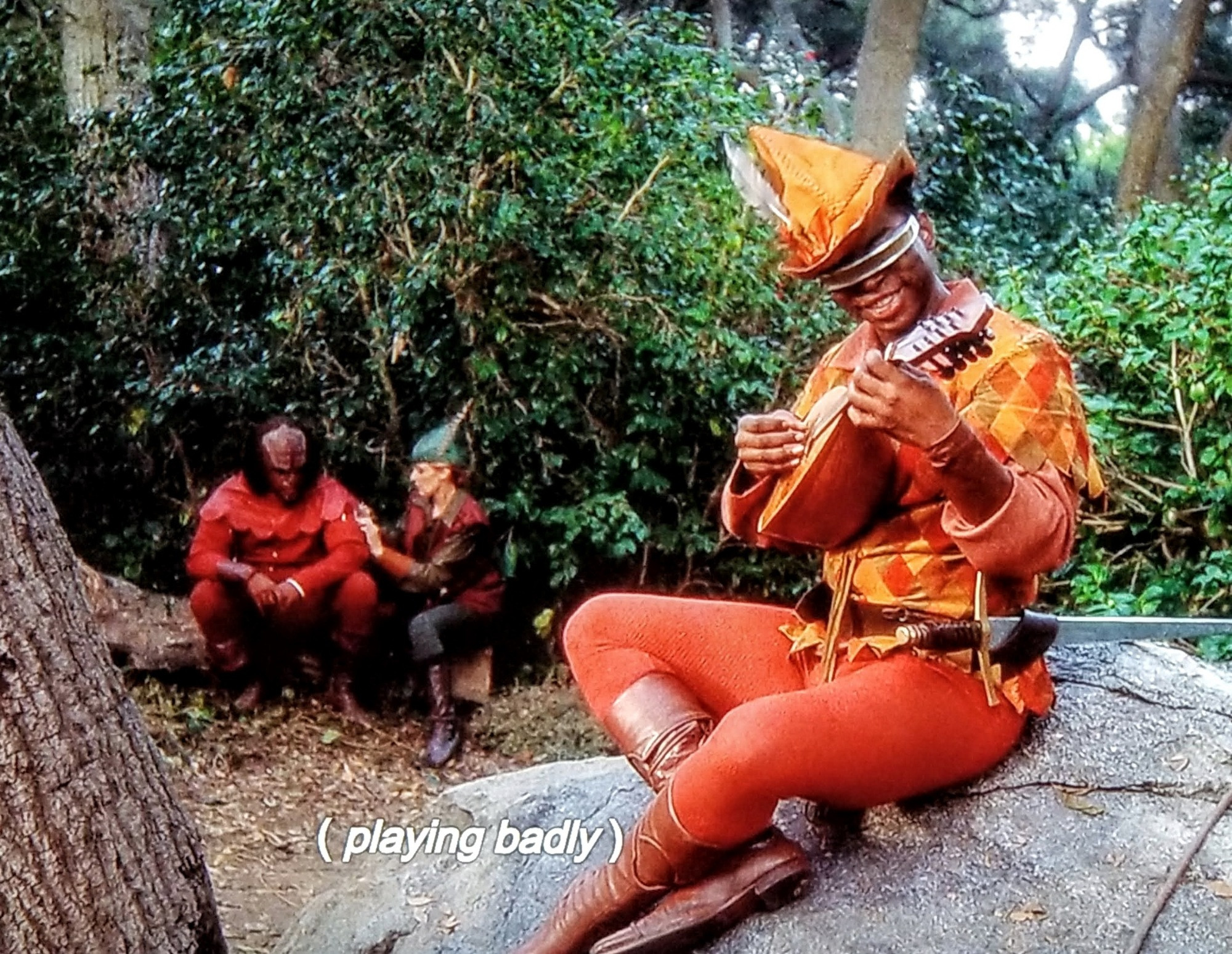 Star Trek The Next Generation scene. Ok, so we're out in a forest and everyone is dressed like they're doing Robin Hood cosplay. In the background is Worf and Crusher. Crusher is tending to Worf's arm, so he probably has an ouchy. Worf, who is sulking, is wearing a red fancy getup, and Crusher's looking Renaissance festivally, with a pheasant feather in her green hat.  Up front is Geordi. He's sitting on a big rock wearing orange tights with sword in the belt loop, in an orange and yellow checkered tunic top and a 3 musketeers shaped feather hat. He's playing a mandolin and smiling big like Leah Brahms just said yes to his friend request on the facebook. Closed caption reads, "(playing badly)"