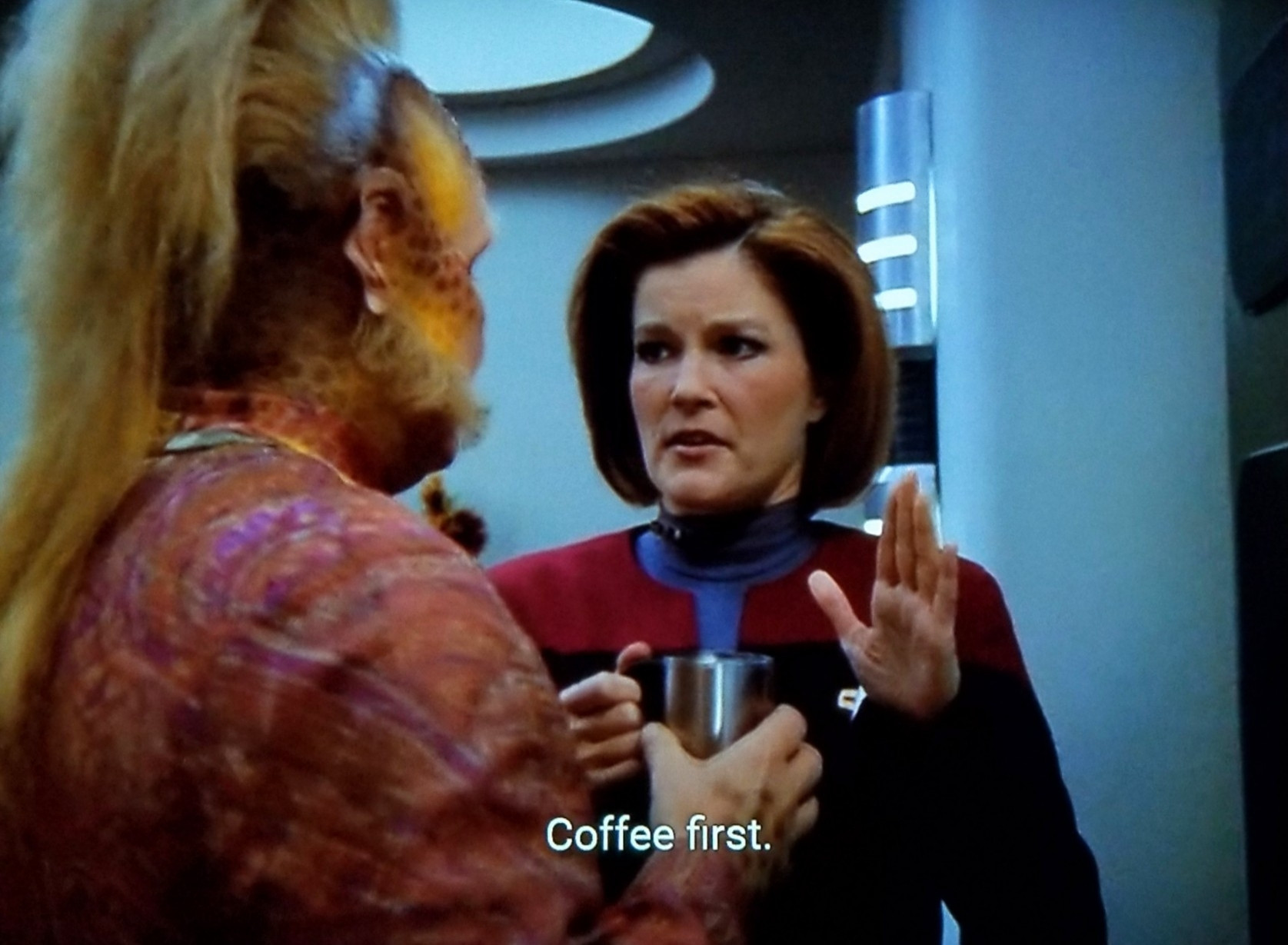 Voyager scene. We're in the kitchen. Neelix is standing and handing off a coffee cup to Janeway who is next to him. She's holding up her other hand in the stop sign hand motion. Closed caption reads, "Coffee first."