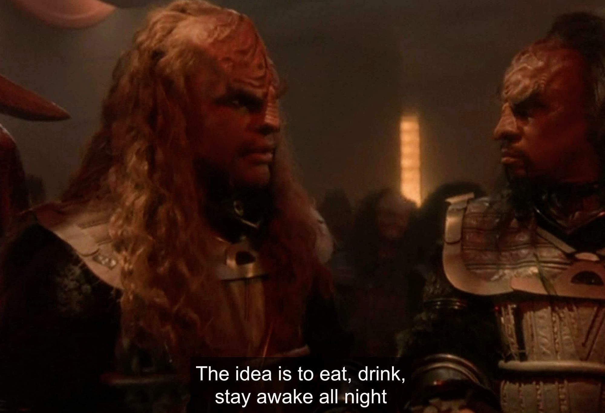 Worf and another Klingon are standing and talking. Both are dressed in traditional Klingon uniforms with silver plated shoulder/chestal areas. Both have long and flowy hair. Both look very serious. It's dimly lit and others are in the background. Closed caption reads, "The idea is to eat, drink, stay awake all night."