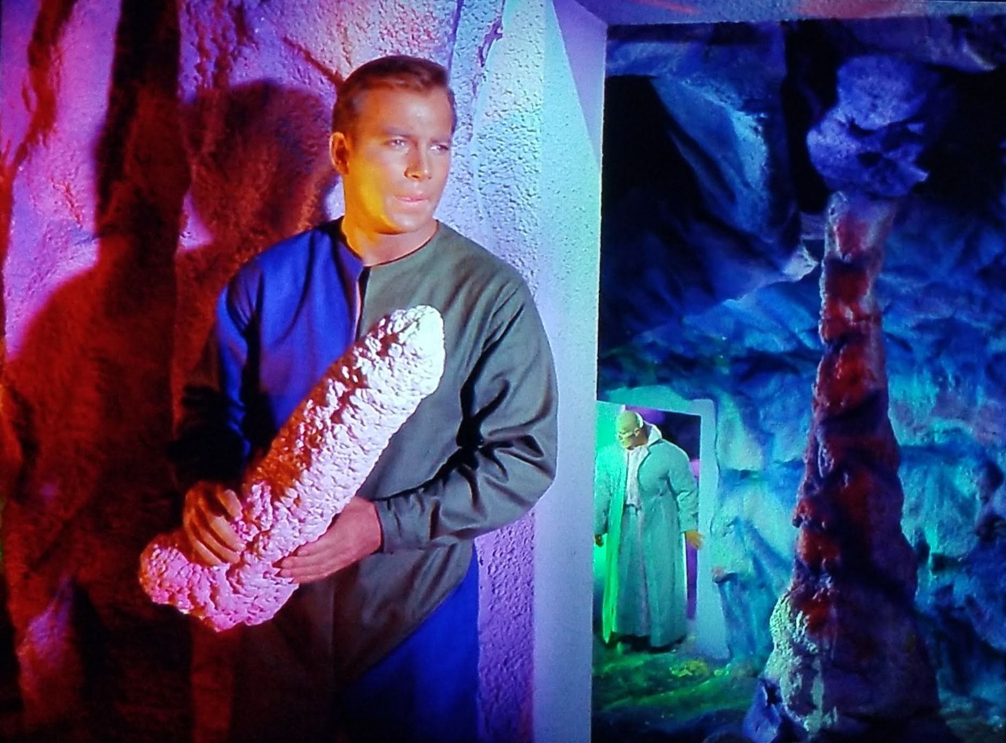 TOS scene. We're in a cave that also has a wall and a far off door carved into it. It's a technicolored cave, lit in purples, reds, blues, and greens. I'm not entirely sure why, but someone was trippin' balls on set that day. Ok, so Kirk is hiding behind a corner up close to the camera, and further away in a different part of the technicolor dream cave is a swol and very tall bald alien. He looks menacing despite wearing a cream green long jacket over a weird flared collared shirt. Back to Kirk, he has a...hmm...so he's holding a large rock to use as a weapon, yeah that's it. It's an oddly long rock though, maybe baseball bat length but with way more girth. And it's pointing upward, with extra "rockage" near the base. (It looks like a dong.)
No caption.