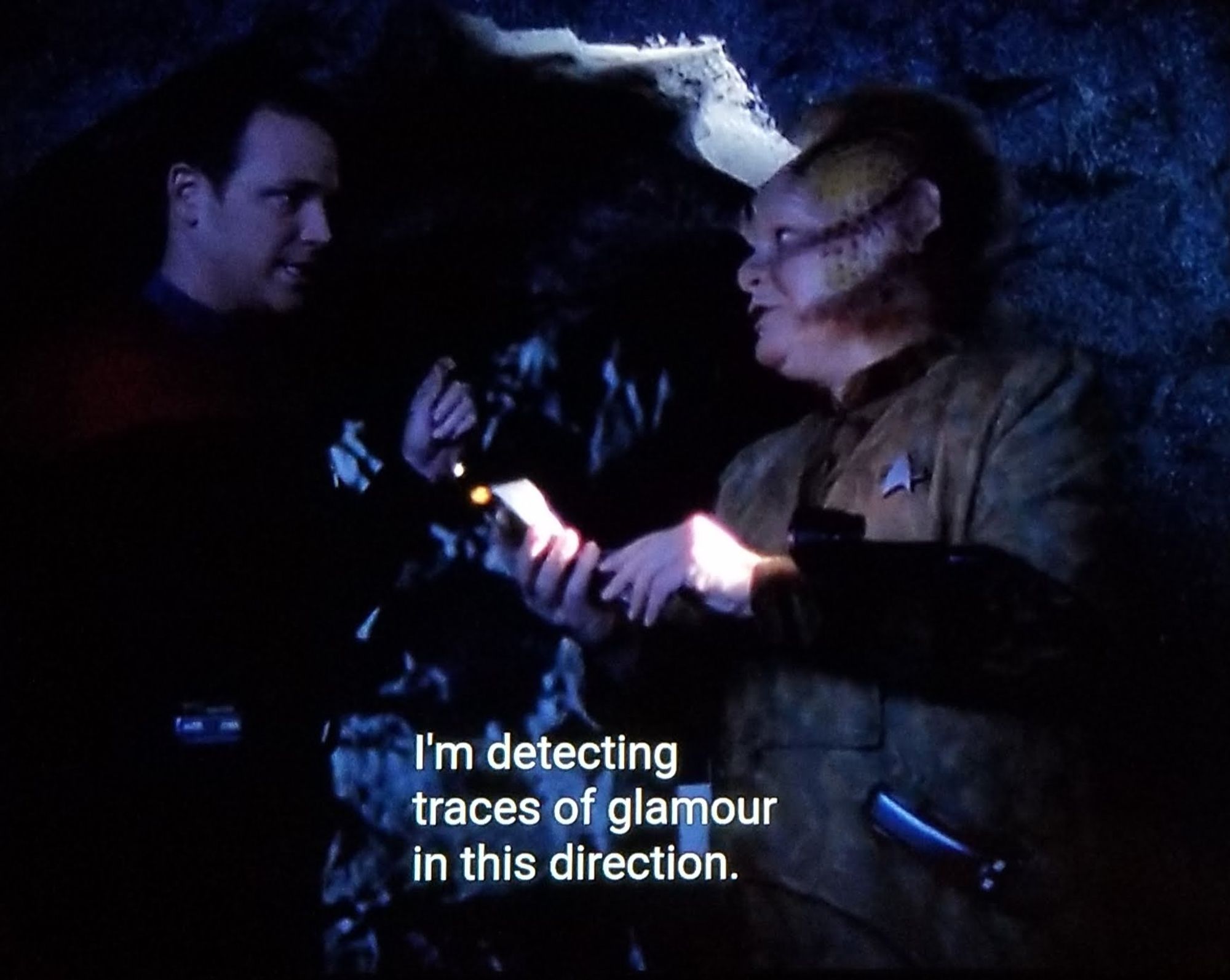 Voyager scene. We're in a dark cave or maybe we're just outside at night? My screen capture sucks so we'll never know. Anyhow, Neelix with his alien face dots and long weird whiskers is holding a tricorder and telling something to Tom Paris who is next to him in this dark weird outdoor cave. Closed caption reads, "I'm detecting traces of glamour in this direction "