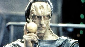 Gul Dukat is pictured. He's a boney humanoid alien fella. Gray skin, with ridges around his eyes, up through his forehead, down his neck and nose, and generally in other places we can but probably shouldn't imagine. He's making a coy sort of here kitty kitty look, and holding a baseball between thumb and forefinger up near his face, looking side eye at someone.