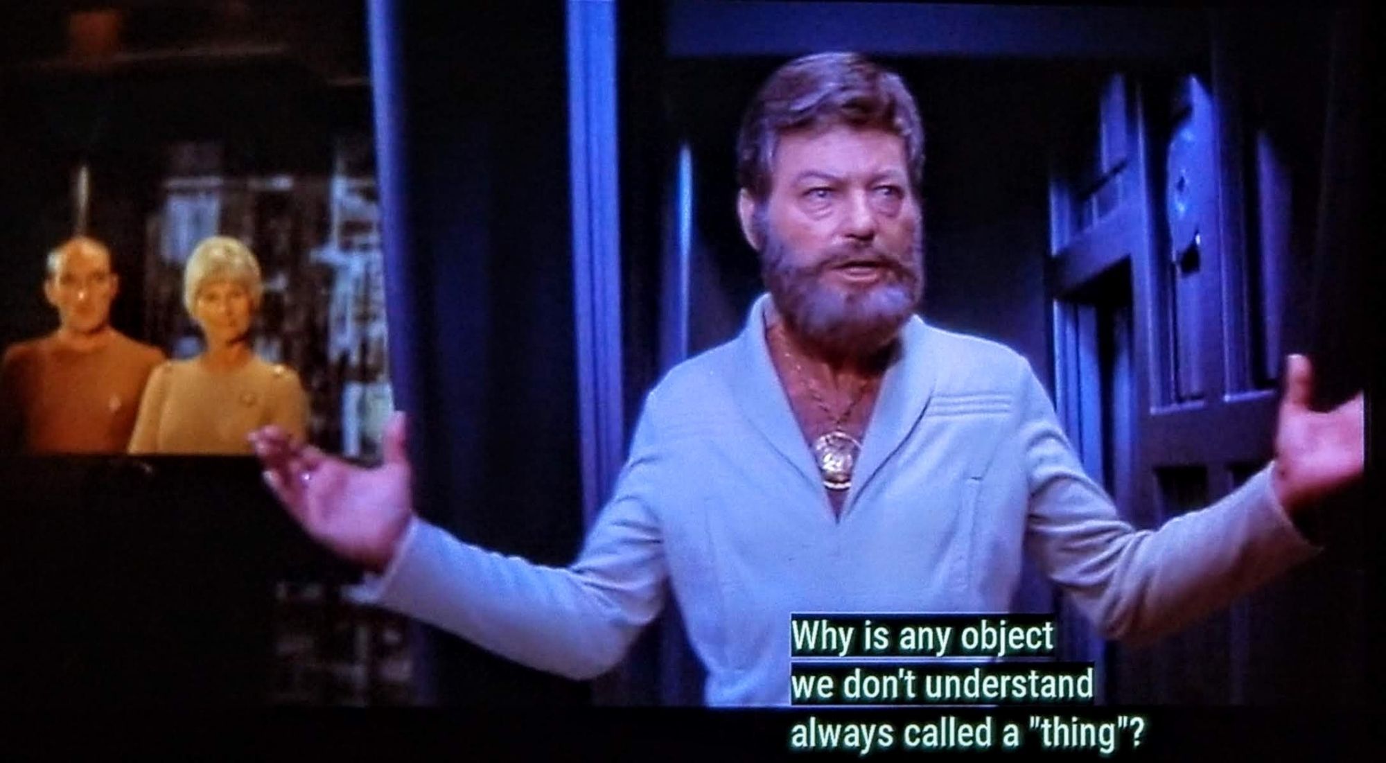 It's a Star Trek The Motion Picture scene! Okay, so here we see Bones McCoy on ship and then 2 onlookers in the background wearing yellow and orange. Bones is wearing a cream colored jumpsuit uniform that was the style in the first movie and then literally never ever seen again. His chestal region is open to an aggressive v-neck that runs up into a big collar. In and amongst his hairy forest of a chest is a gold medallion, similar to those worn on the Barry Gibb talk show. He has a large and buoyant beard, kind of like the Dad in Teen Wolf when he (spoiler alert) also turns into a wolf. Bones is holding his hands out in a exasperated good grief kind of motion. Closed caption reads, "Why is any object we don't understand always called a "thing?"