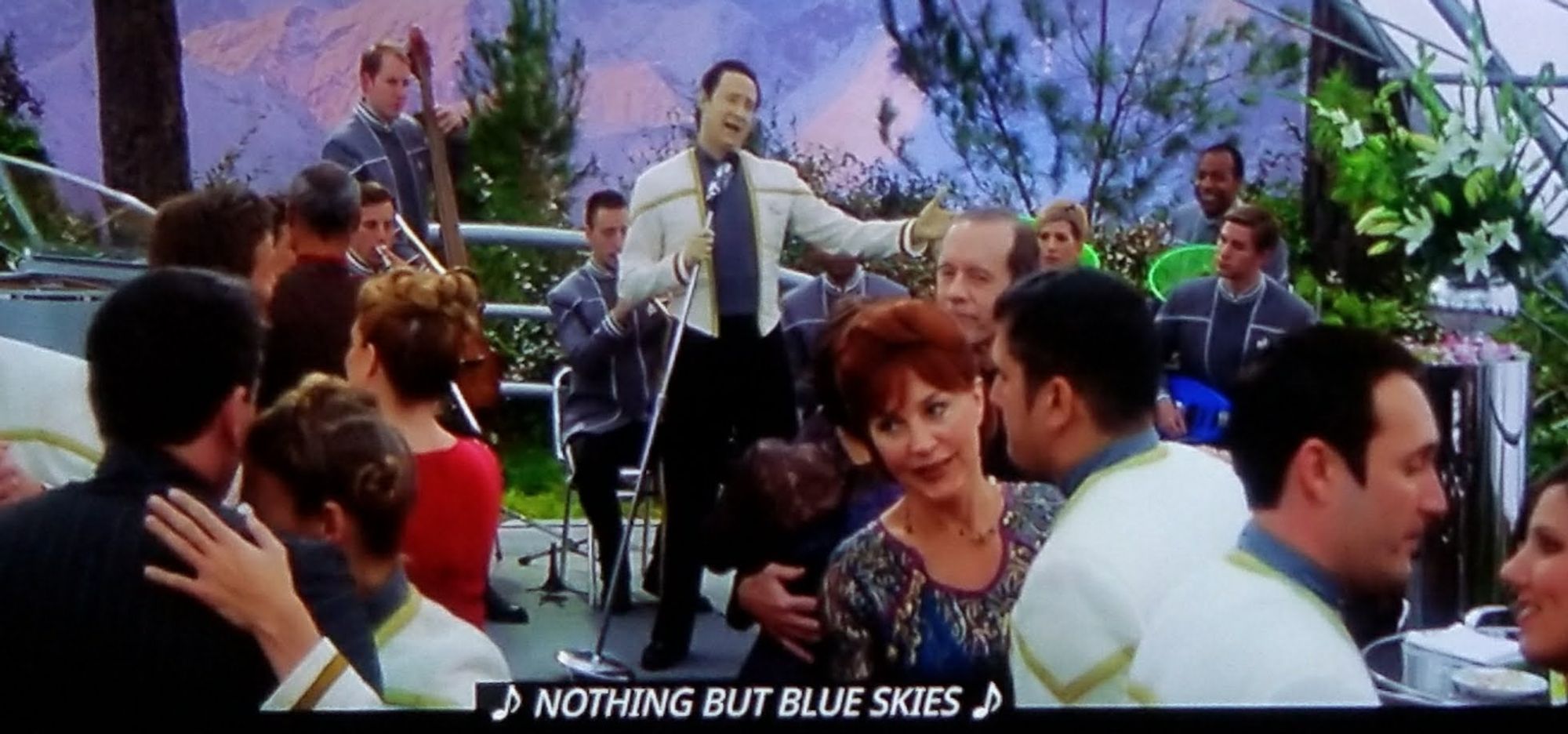 TNG movie. We're having a fancy party, starfleet folks in white dress uniforms are dancing with others in party civilian clothes. There's maybe mountains in the background and definitely some trees. Beyond the dancing couples is a big band playing music, and Data is standing in front, holding a microphone stand at an angle and singing. He's holding out an arm and really giving them the ol' razzle dazzle. Closed caption reads, "🎵 NOTHING BUT BLUE SKIES🎵"